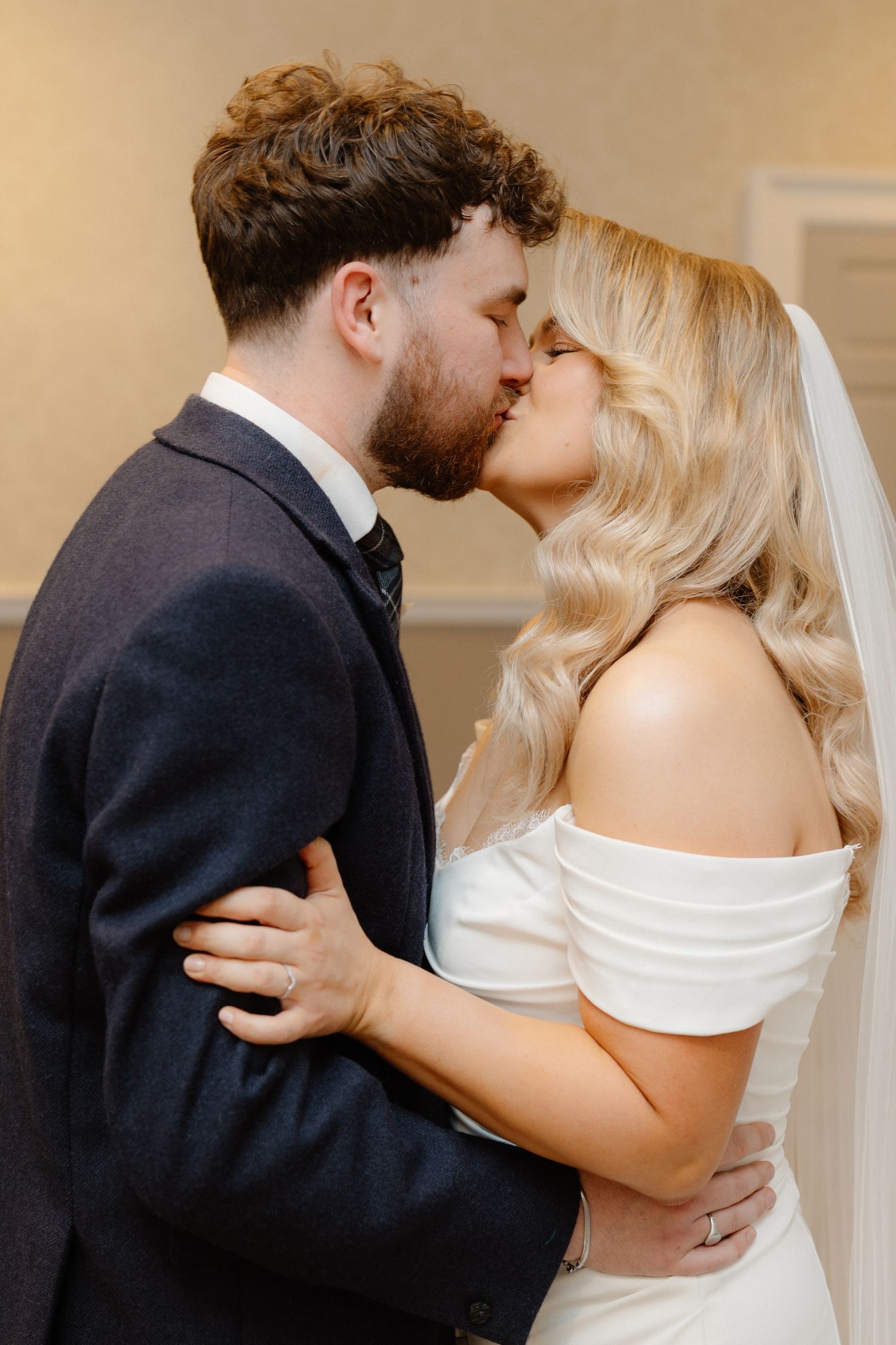 A bride and groom share a romantic kiss, she wearing an off-shoulder white gown with veil, he in a navy suit.