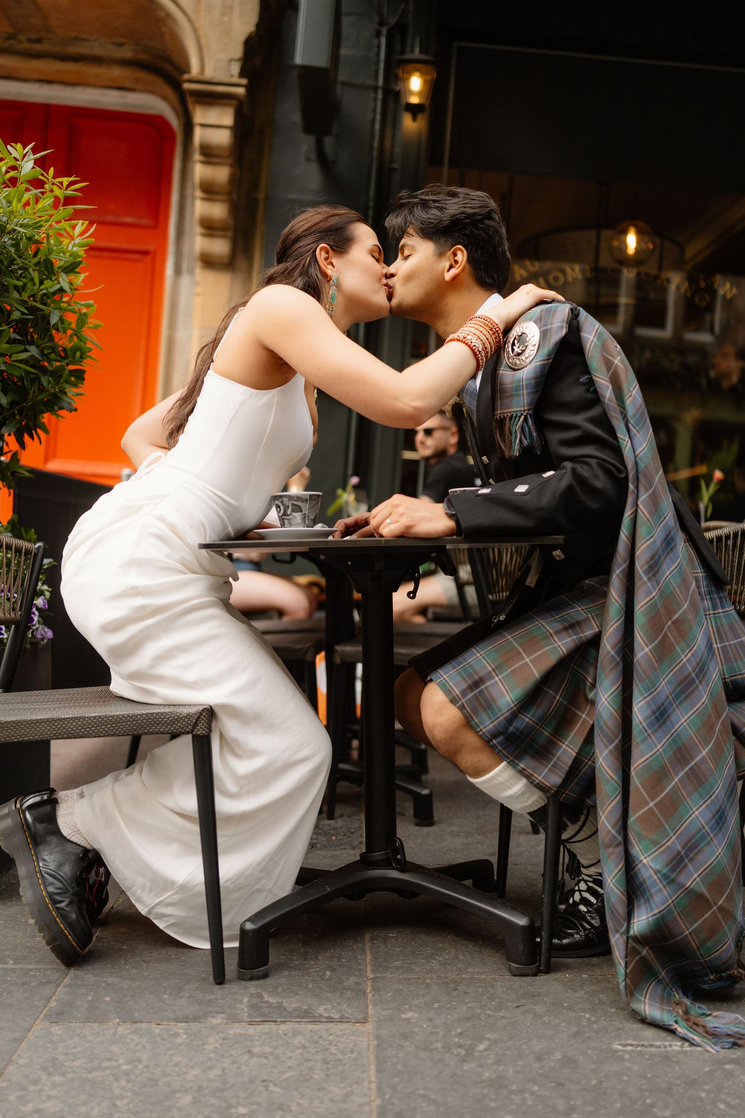 Couple sharing a kiss at an outdoor cafe table, bride in white dress and groom in traditional Scottish kilt attire.