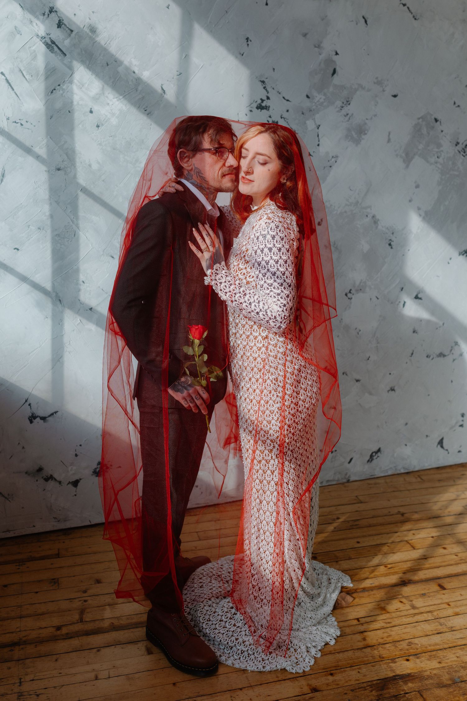 Bride and groom share romantic moment under red veil, she wears lace dress holding rose in sunlit studio.
