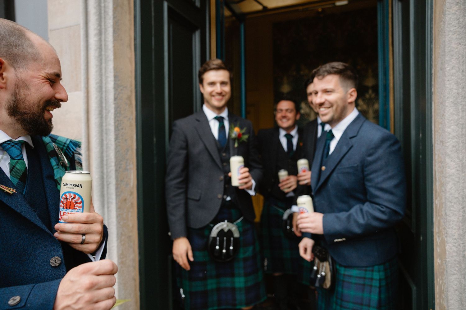 Groomsmen in kilts and suits celebrating with champagne bottles outside a wedding venue.