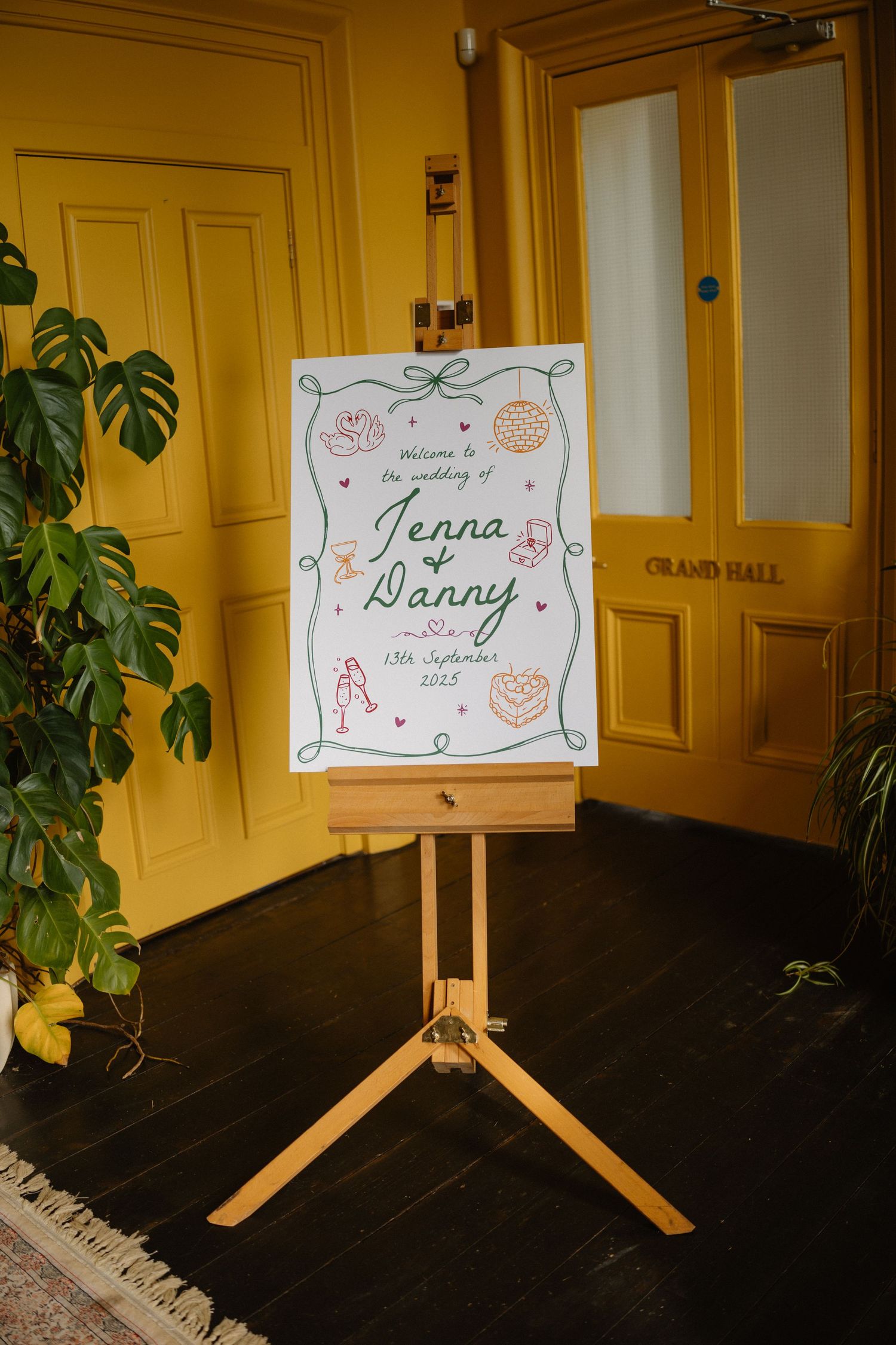 Elegant wedding welcome sign for Jenna and Danny on a wooden easel in front of yellow doors at Grand Hall.