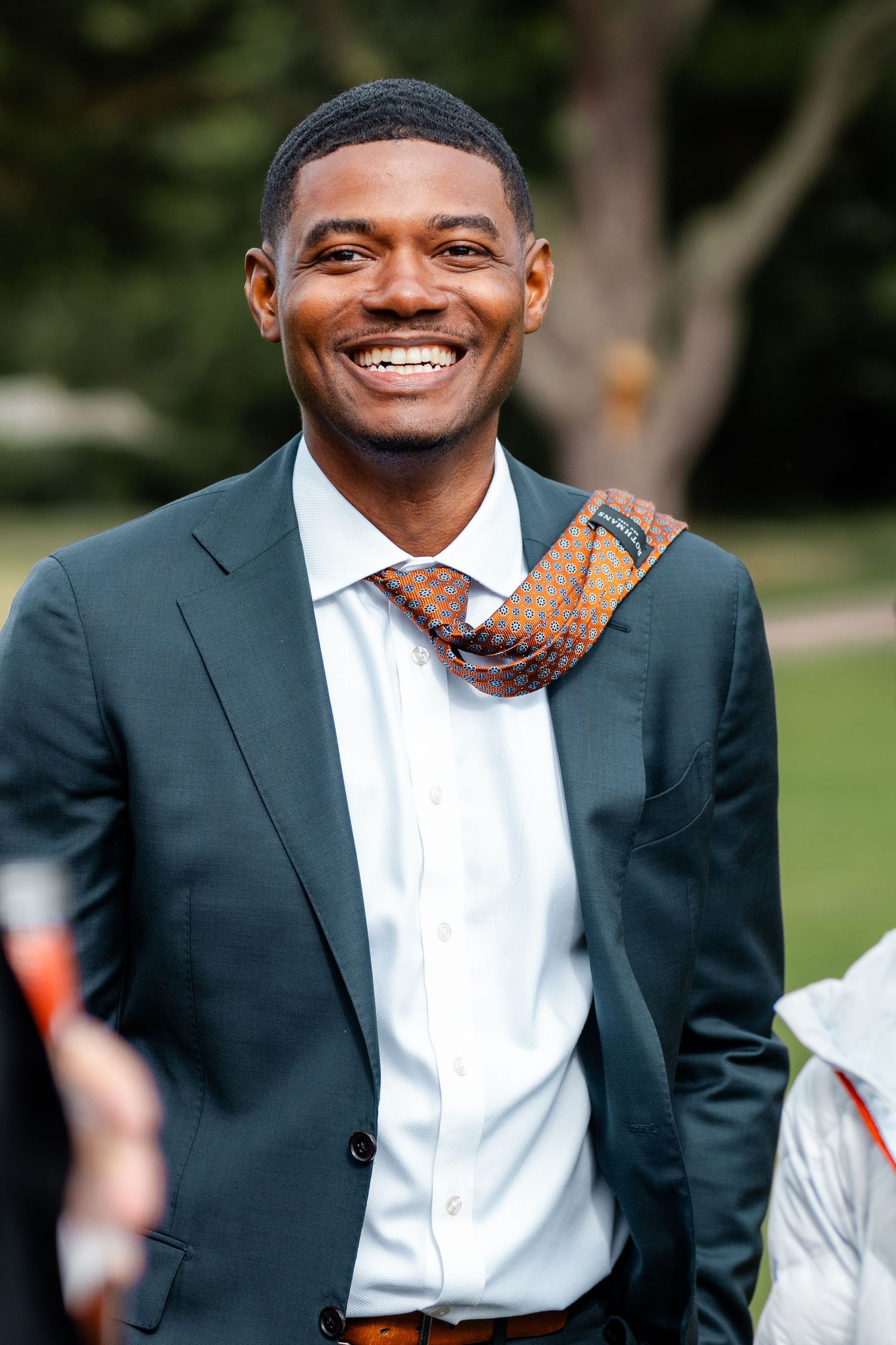 Smiling man in dark suit and orange patterned tie enjoys outdoor wedding celebration in garden setting.