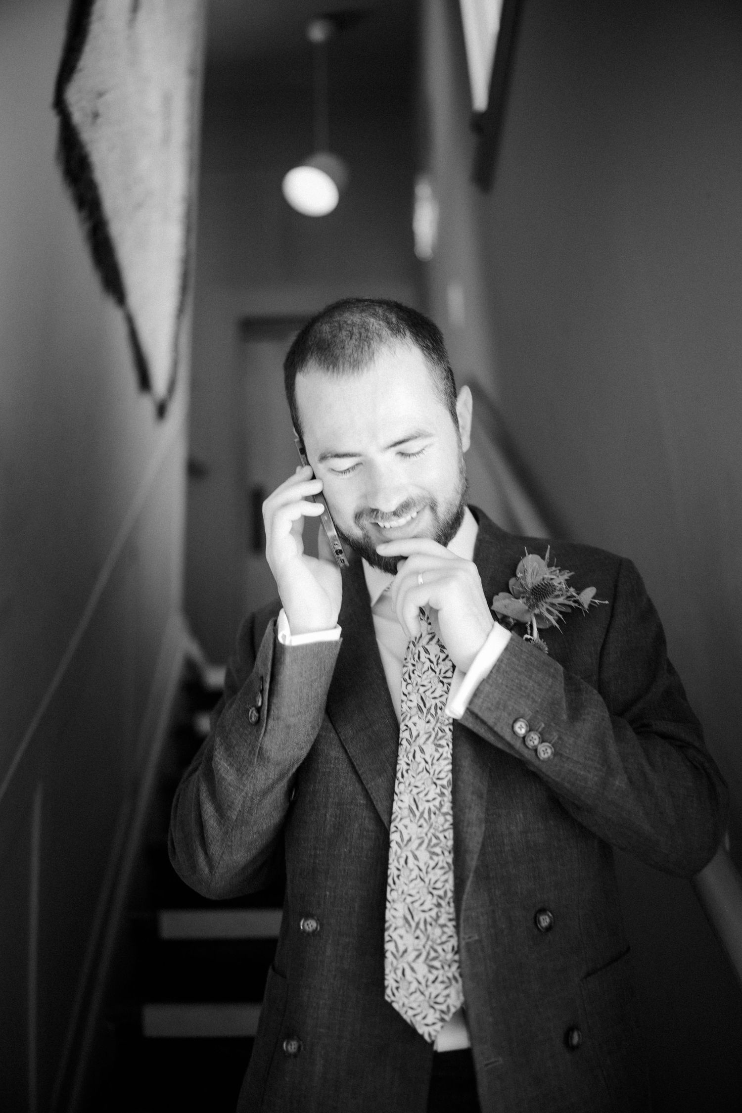 Black and white photo of a groom in a suit with boutonniere, smiling while talking on phone near staircase.