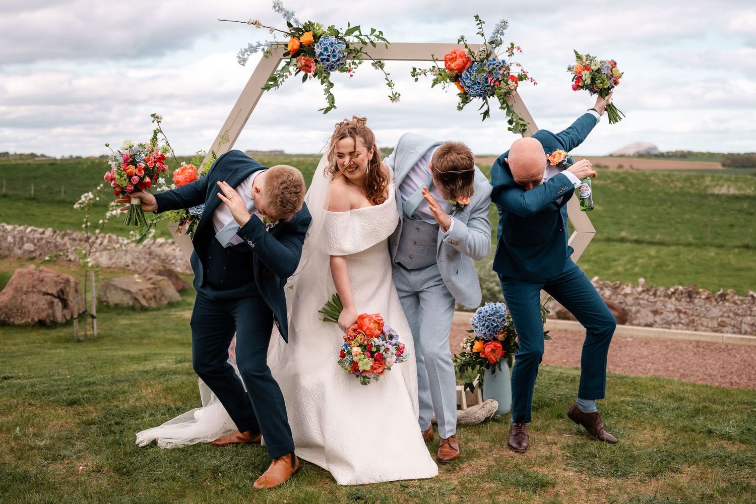 Bride and groom celebrate with wedding party under floral arch, guests striking fun poses outdoors.