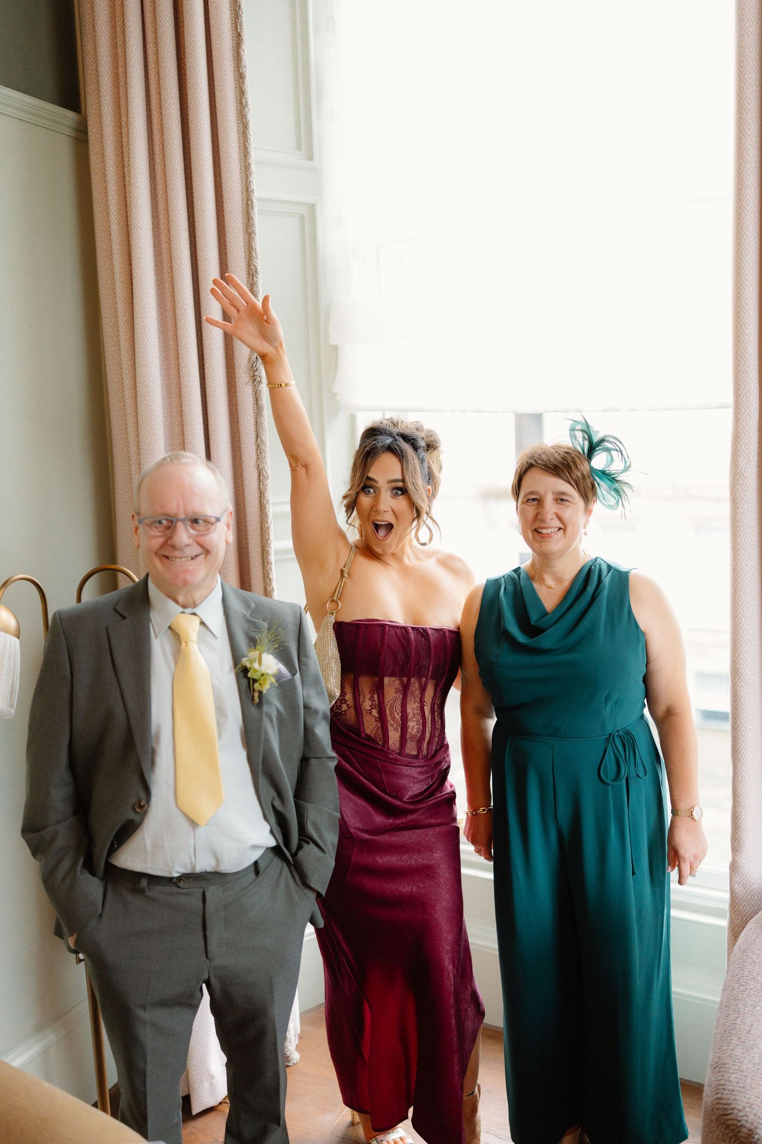 Three wedding guests posing indoors, woman in burgundy corset dress raising hand excitedly between two smiling companions.