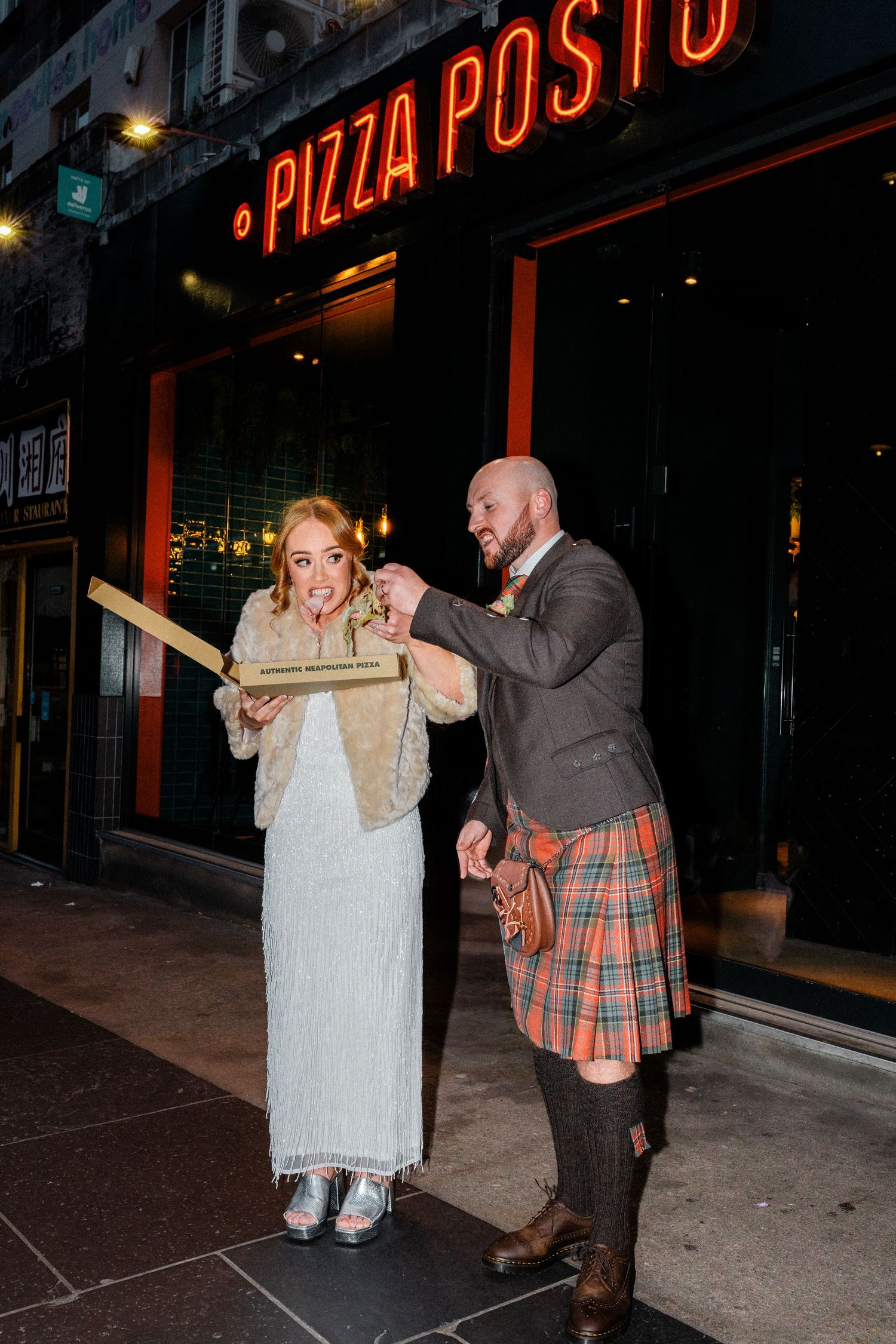 Bride and groom in tartan kilt eating pizza outside Pizza Posto restaurant at night.