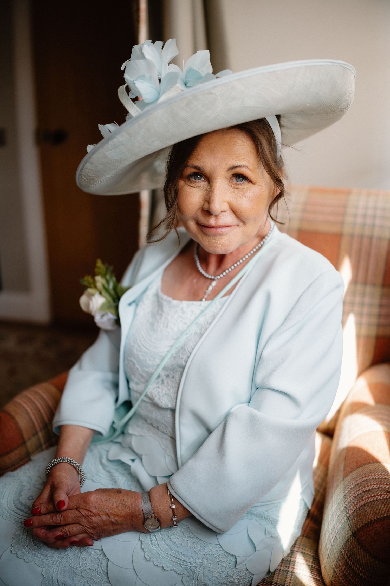 Elegant older woman wearing a light blue wide-brim hat and matching jacket seated in a chair for a wedding.