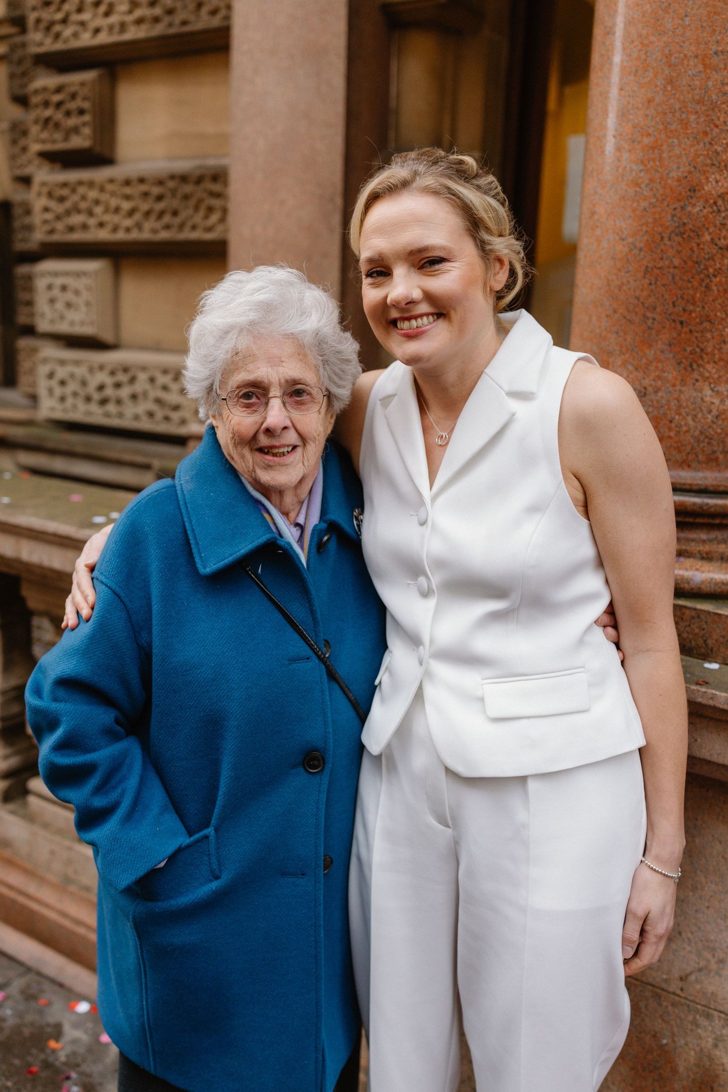 Elderly woman in blue coat poses with young blonde woman in white suit outside a building.