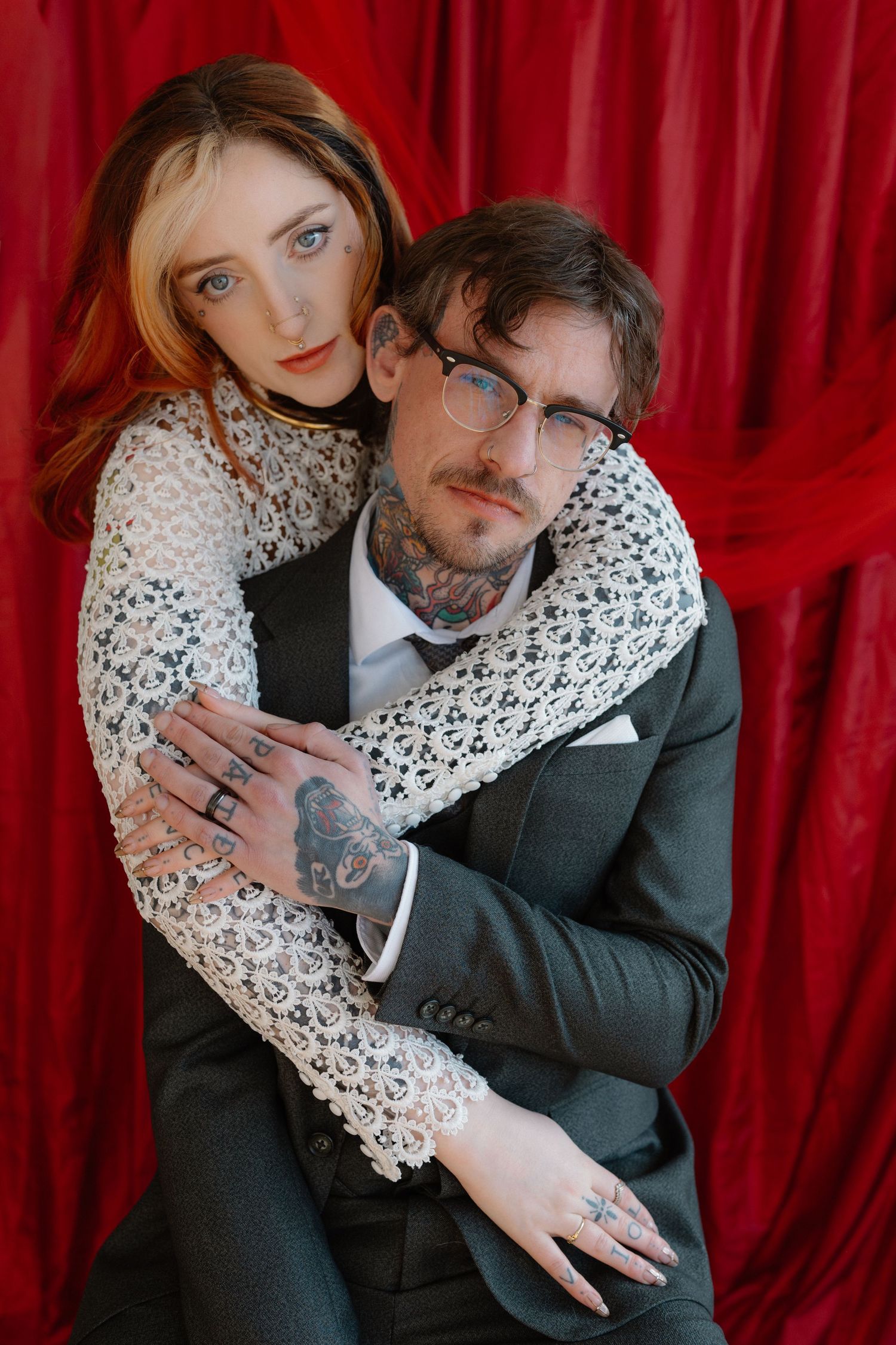 A tattooed couple in formal wear embraces against a red velvet backdrop, the woman in lace hugging a suited man with glasses.
