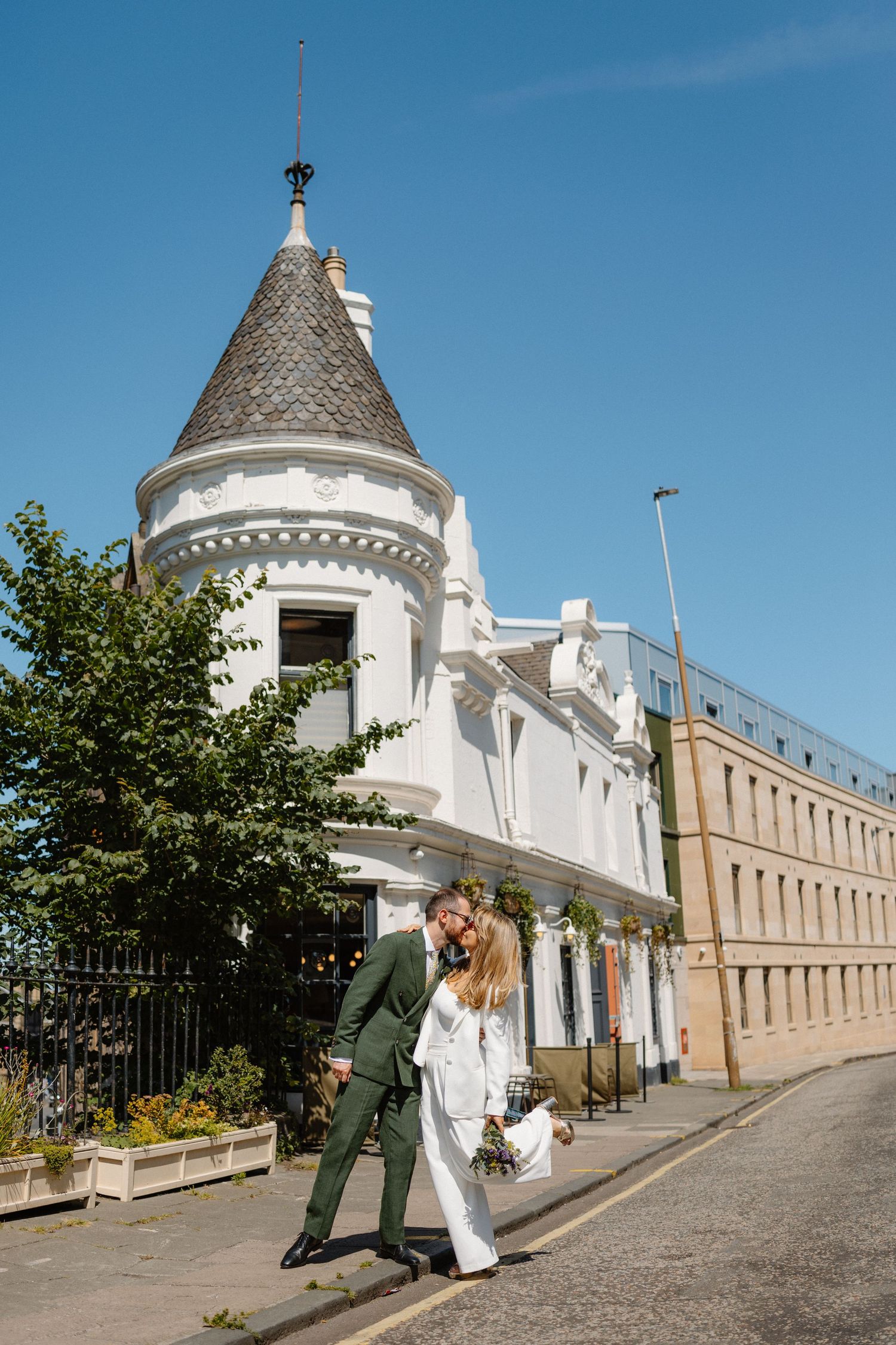 Bride and groom walking outside a white Victorian building with a turret on a sunny day.