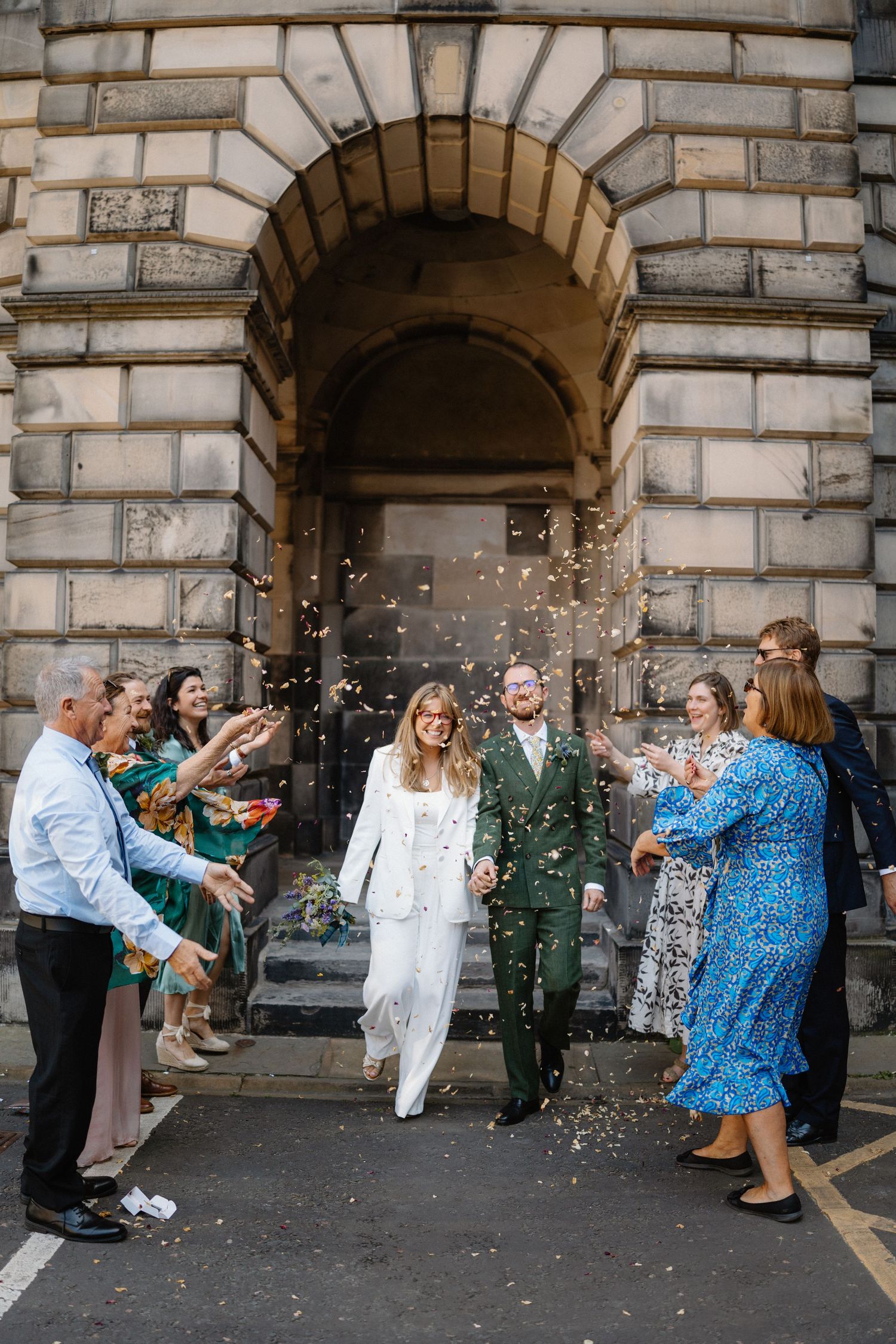 Newlyweds exit a grand stone building as guests shower them with confetti after their wedding ceremony.