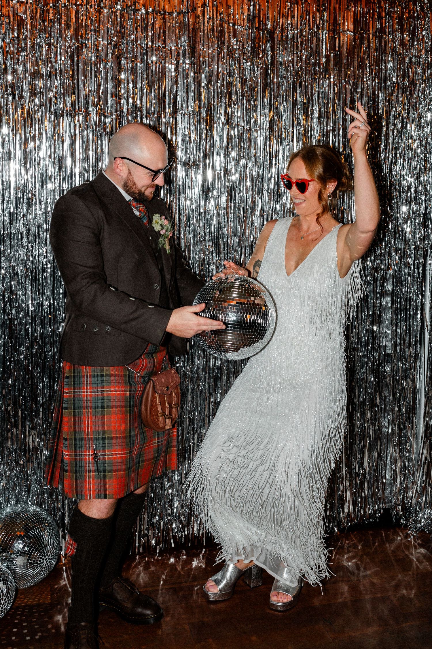 Bride in silver fringe dress and groom in red tartan kilt holding disco ball at wedding reception.
