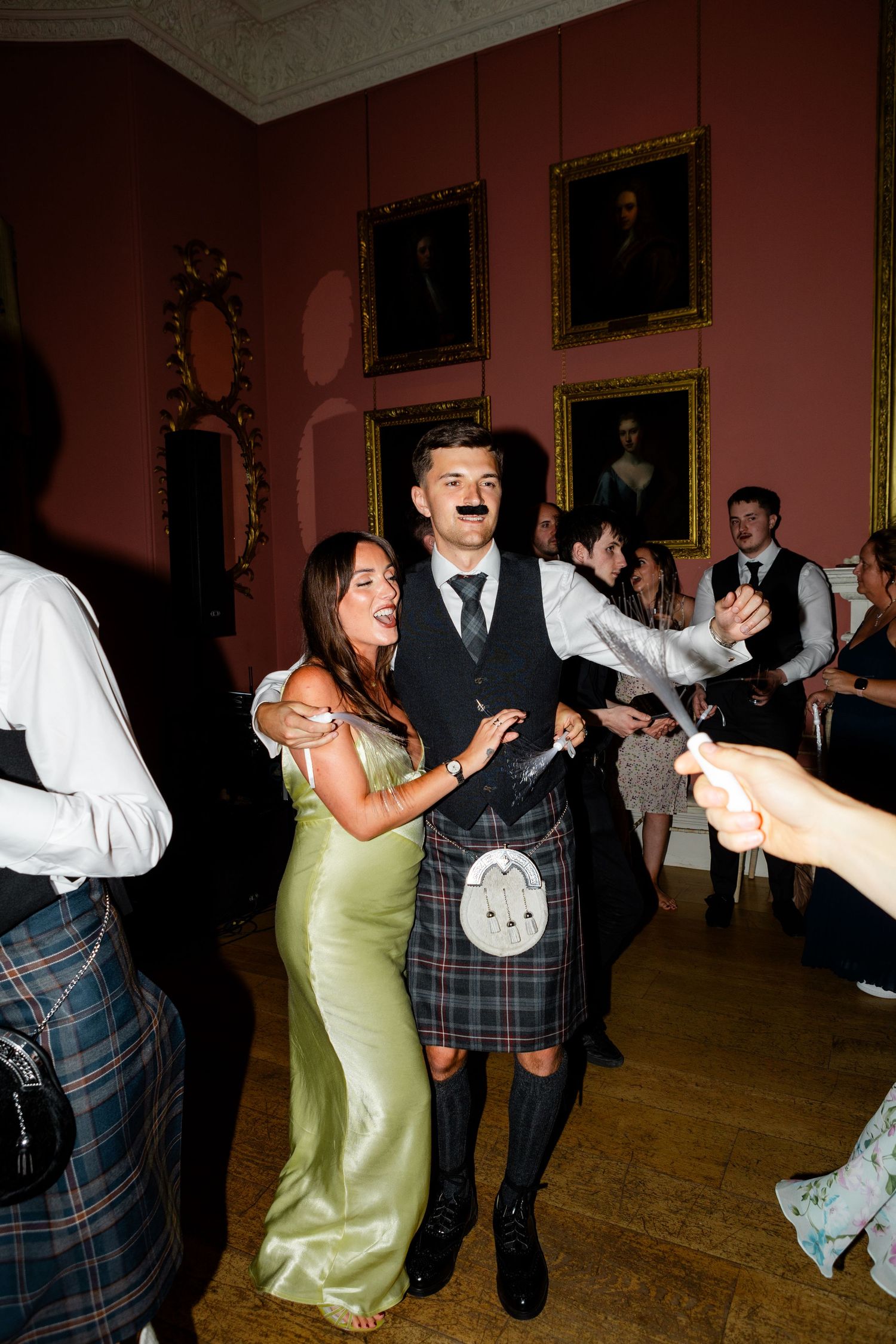 Wedding guests dancing in red-walled Scottish venue, man wearing kilt and fake mustache with woman in green satin dress.