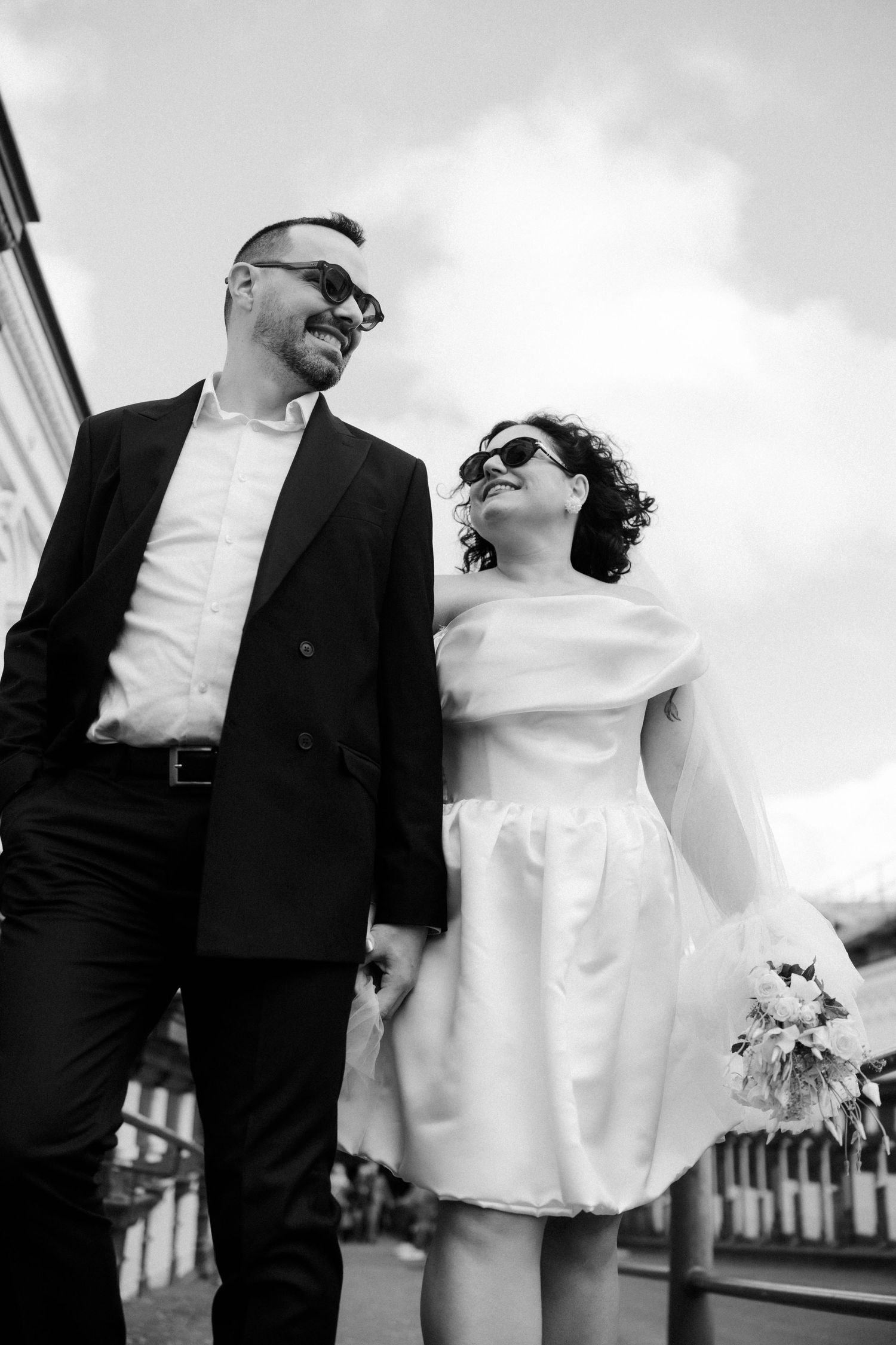 Black and white photo of smiling newlyweds in sunglasses walking together, bride holding bouquet outdoors.