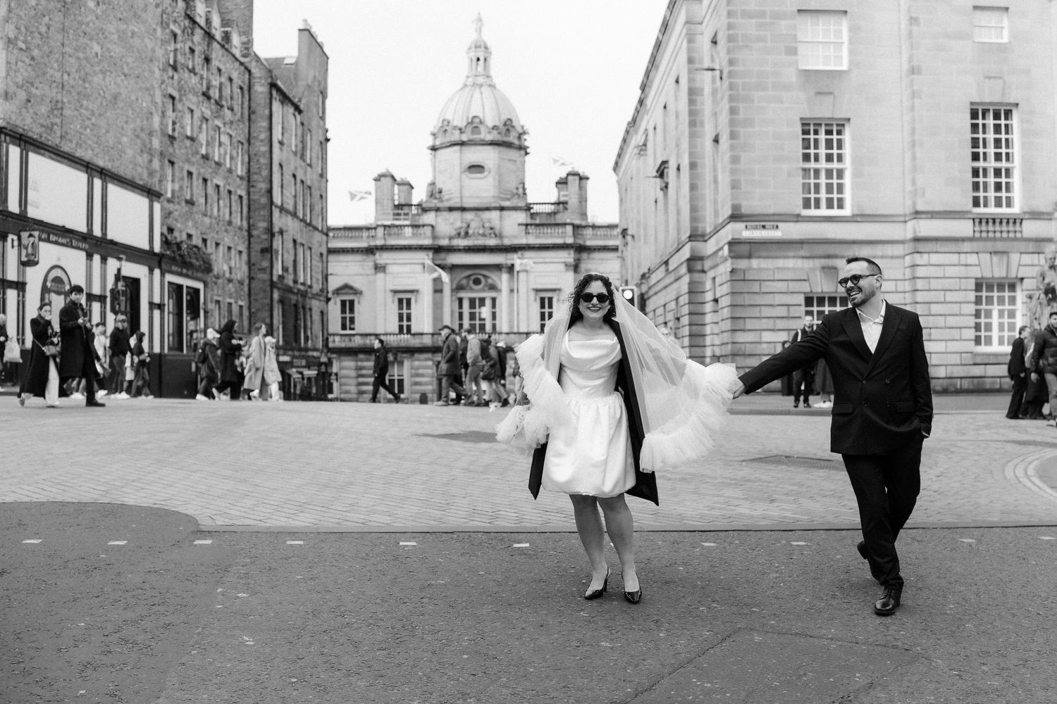 Bride and groom walking through historic Edinburgh city square, bride in short white dress and veil.