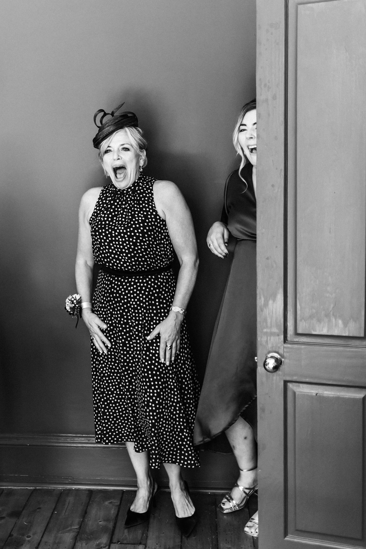 Two women in elegant dresses laugh and pose dramatically in a black and white photo near a door.