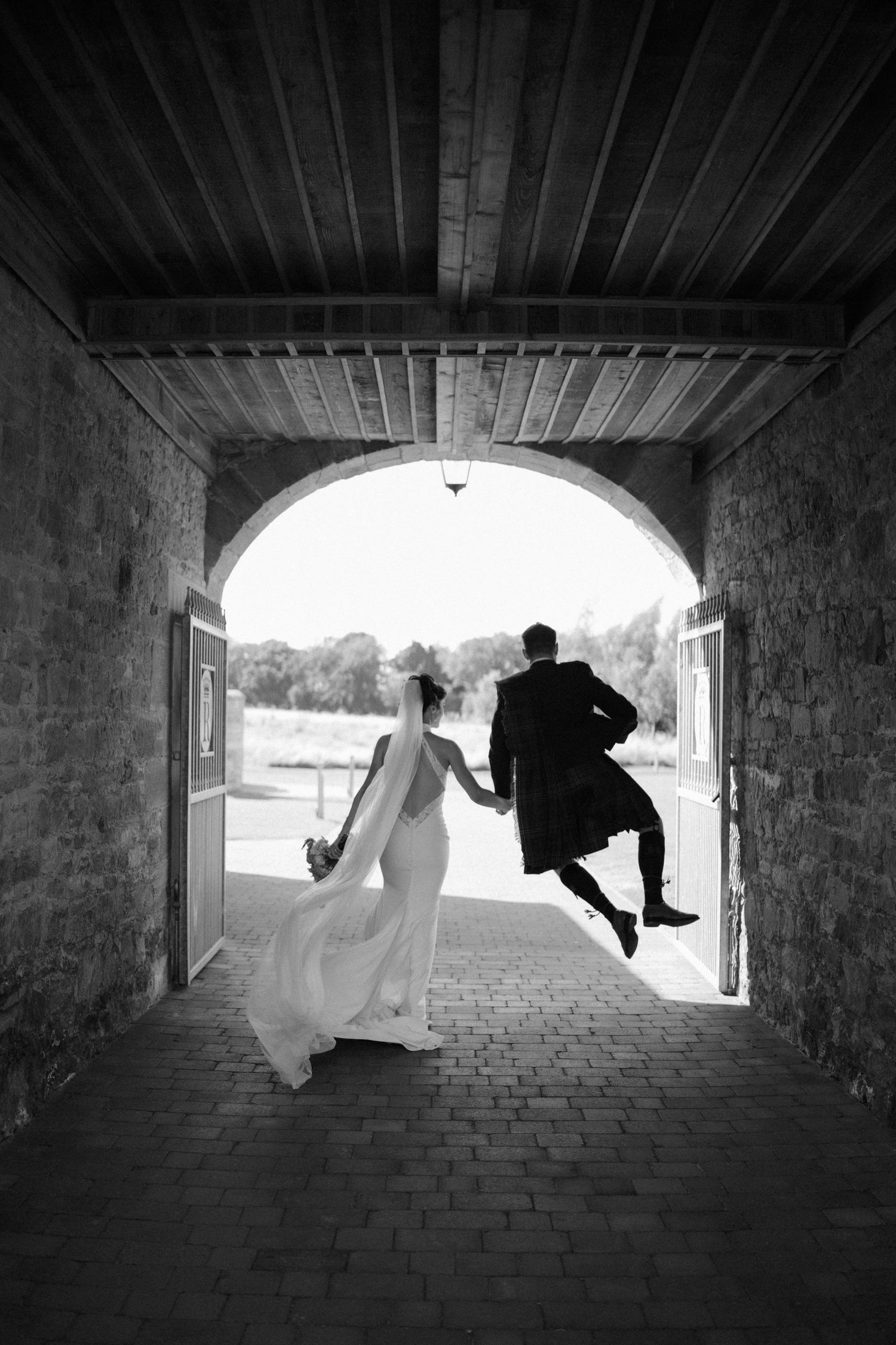 Black and white photo of bride and groom jumping joyfully through an arched stone doorway at wedding.