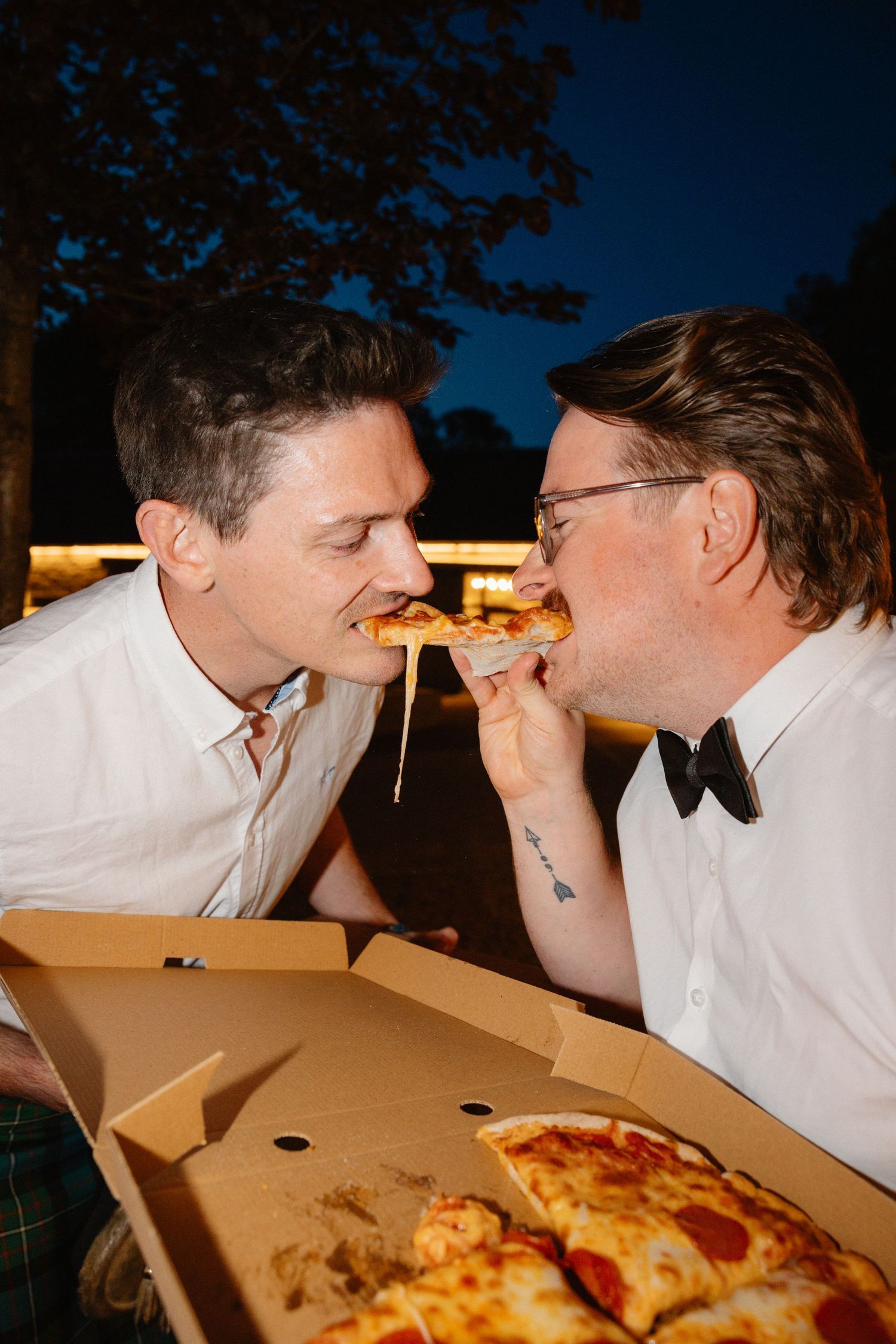 Two grooms share a romantic bite of pizza together outdoors at night during wedding celebration.