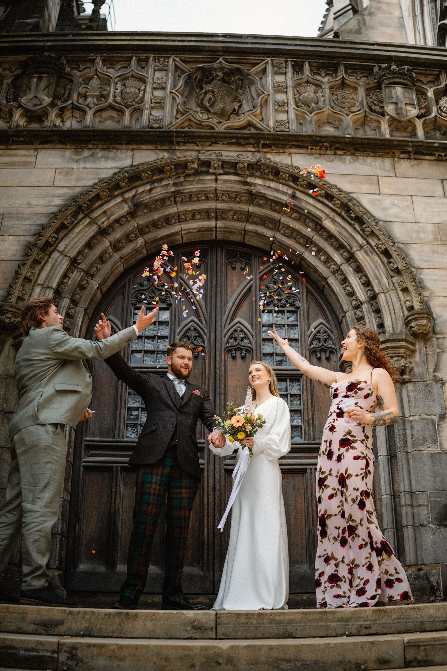 Wedding party throws colorful confetti outside ornate Gothic church doors in Edinburgh.