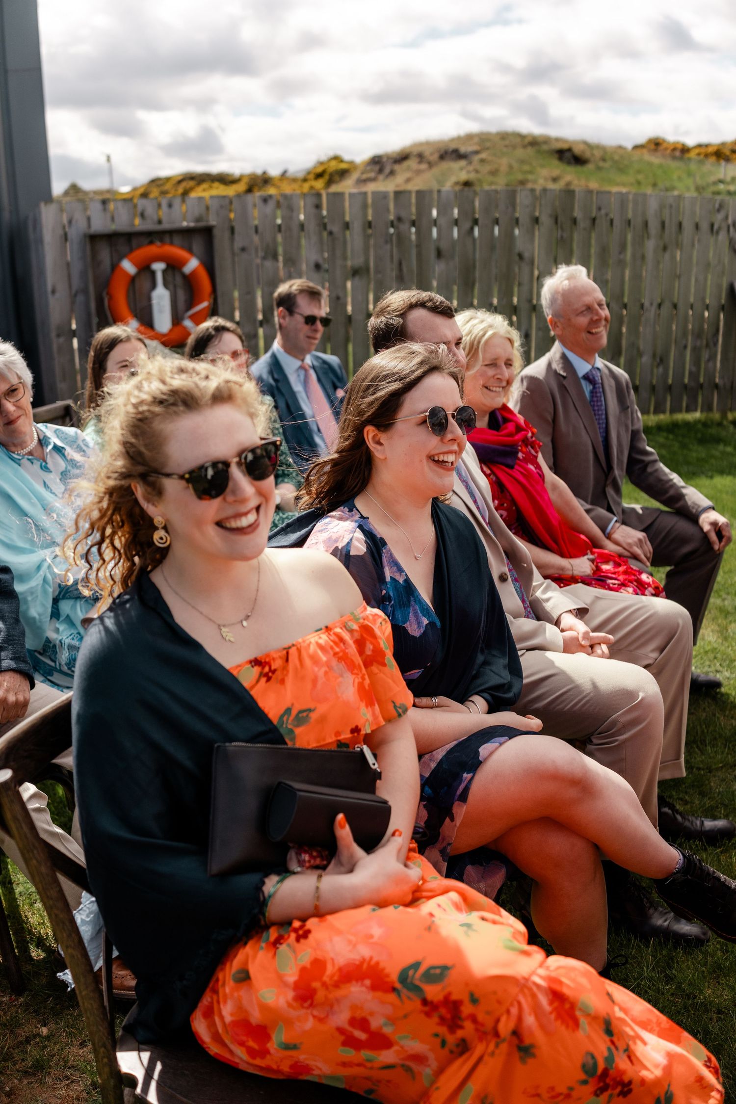 Wedding guests in colorful attire enjoying the ceremony on a windy outdoor day.