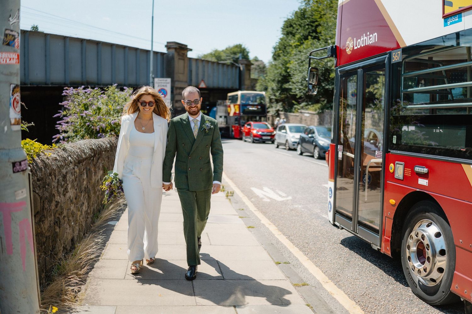 A couple walking on a sunny street with red buses in the background.