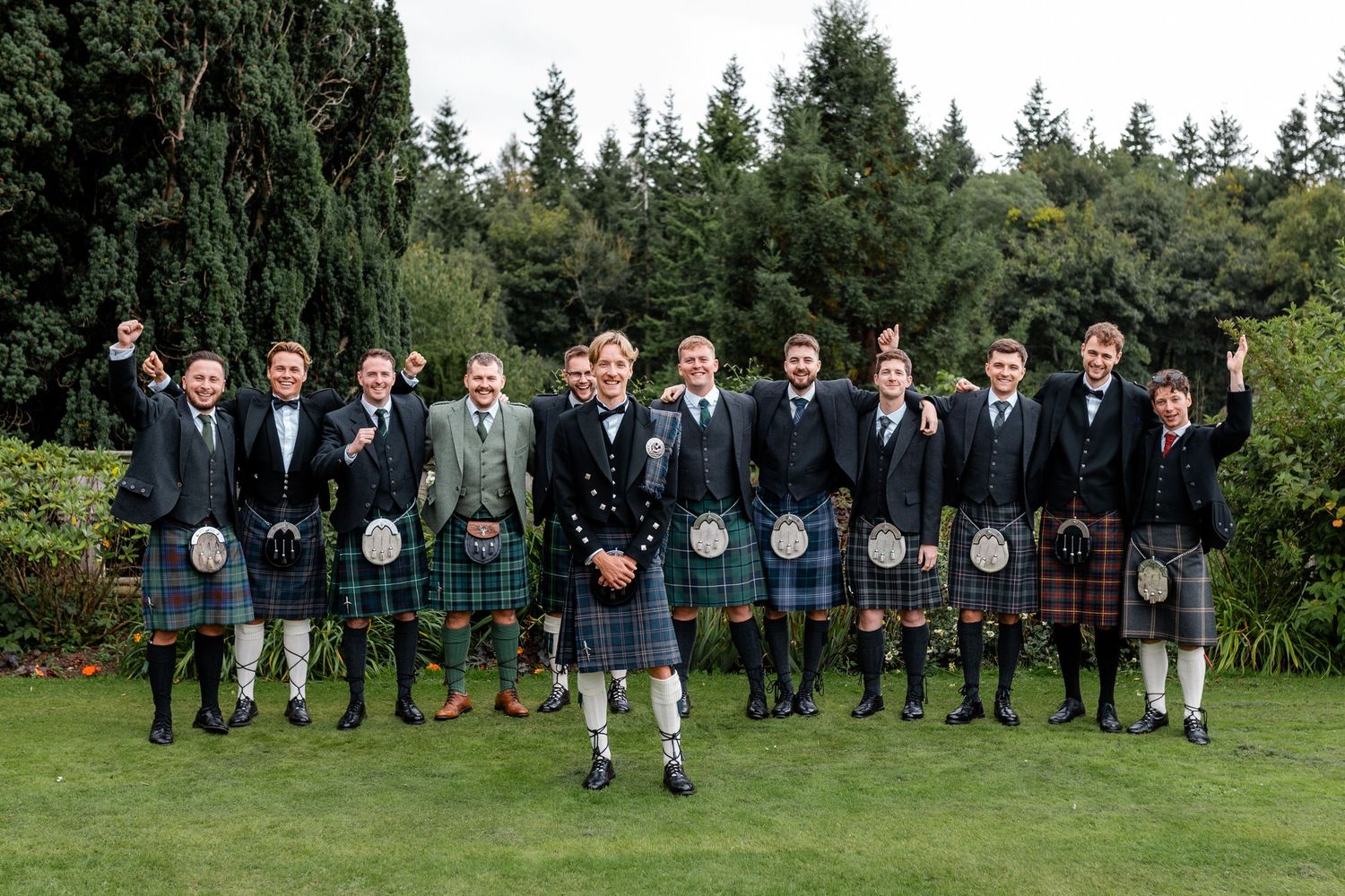 Large wedding party in kilts and formal attire poses in garden setting.