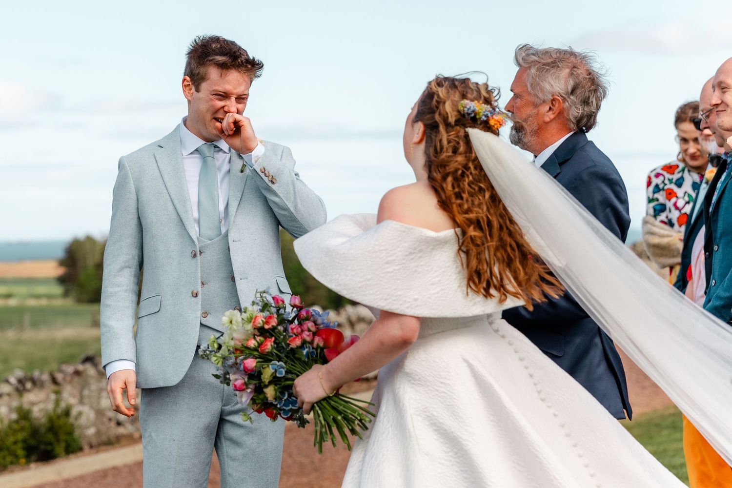 Emotional wedding moment captured outdoors with floral bouquet and flowing veil.