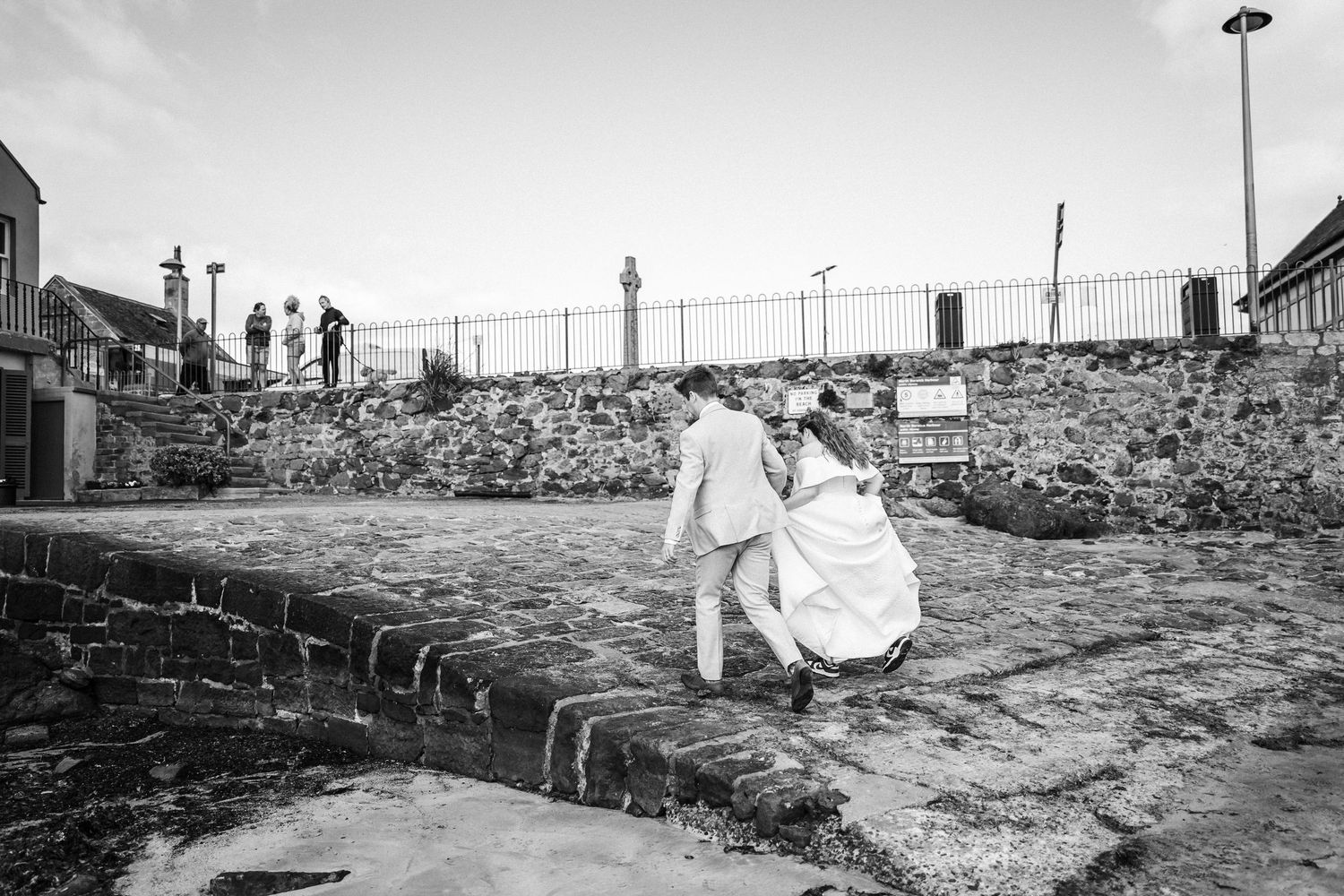 A couple poses for wedding photos on a rustic stone wall with a coastal landscape in the background.