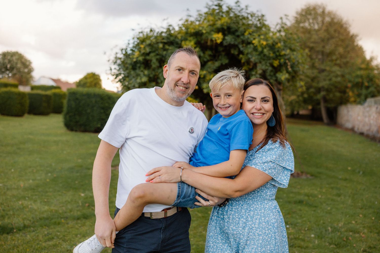 Family of three posing together in garden setting with hedges.