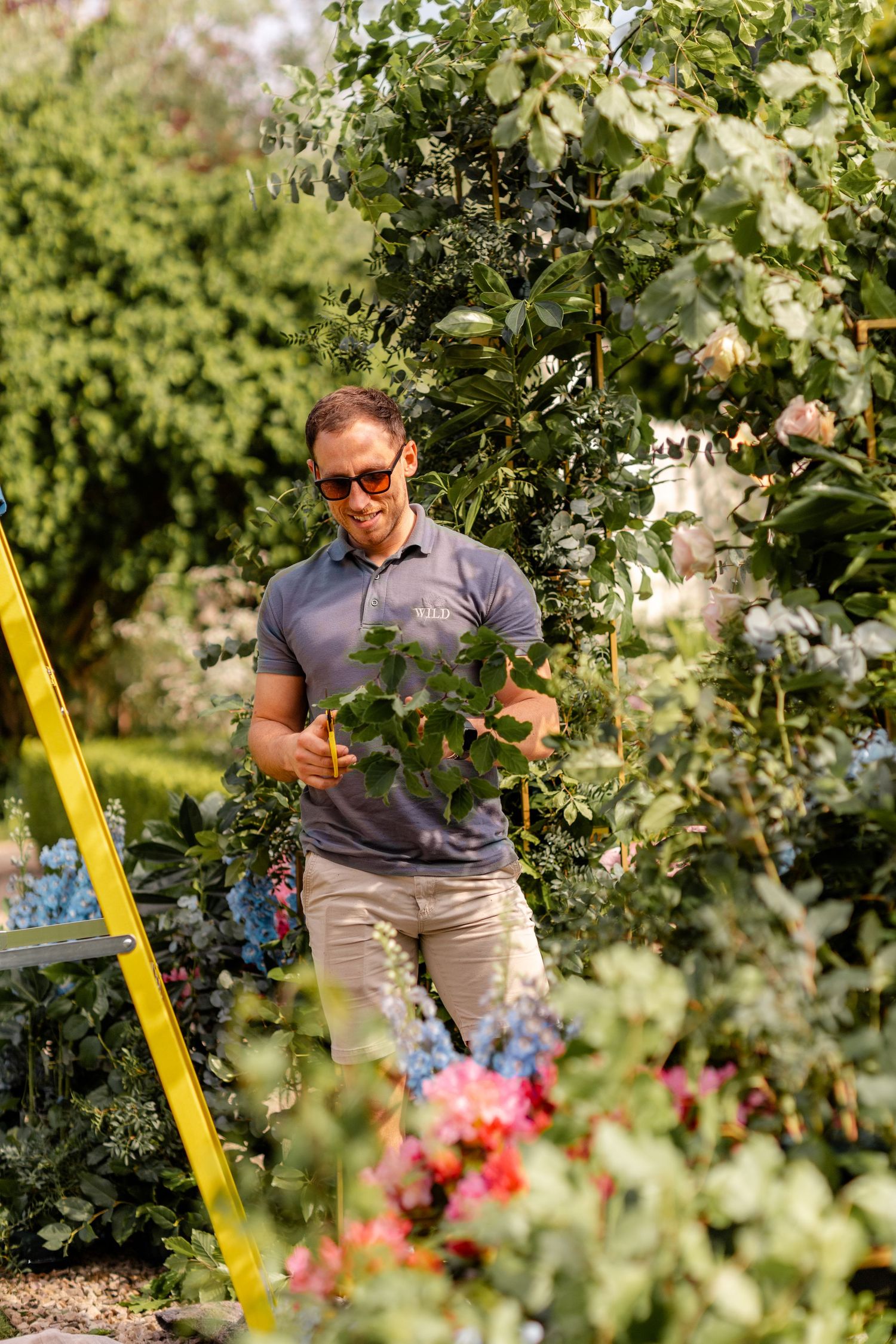 People tending to plants and flowers in a garden with a yellow ladder visible among the greenery.