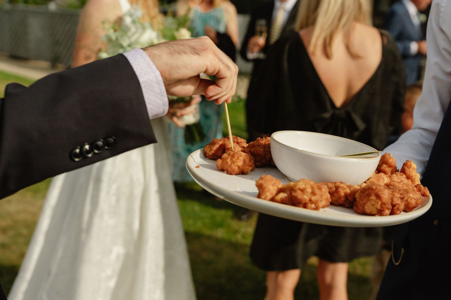 A guest picks up a crispy fried appetizer from a white plate with dipping sauce at an outdoor wedding reception.