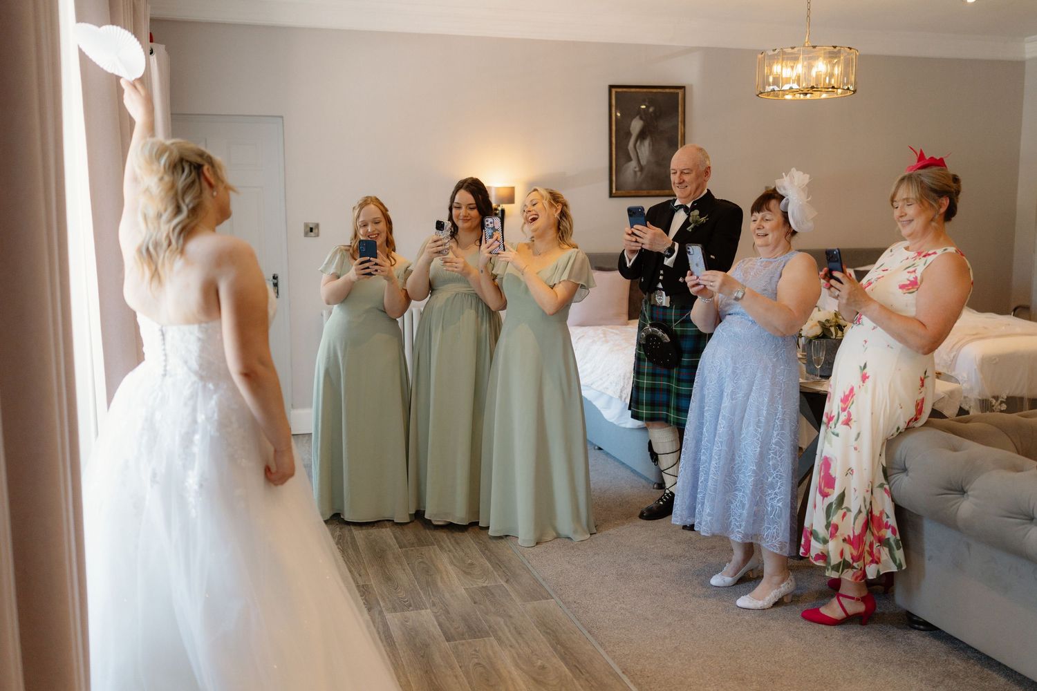 Bridesmaids in sage green dresses react with joy as bride reveals her wedding gown in a bright hotel room.
