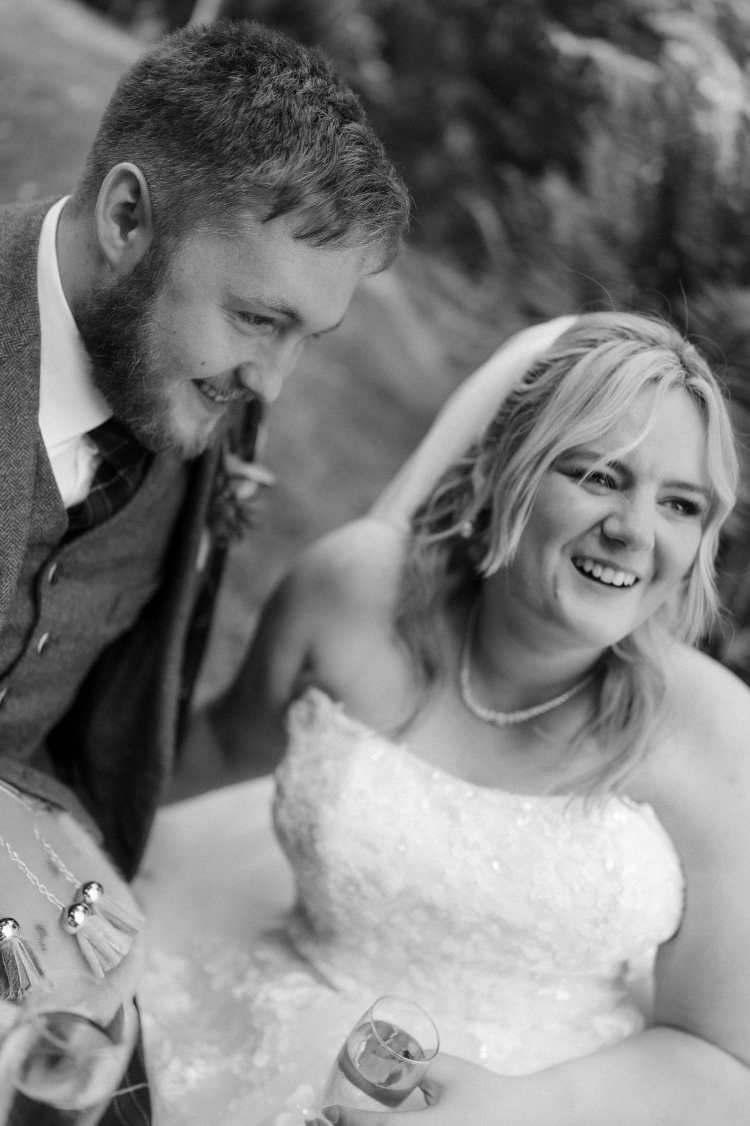 Black and white photo of a laughing bride in white dress and bearded groom in suit at wedding reception.