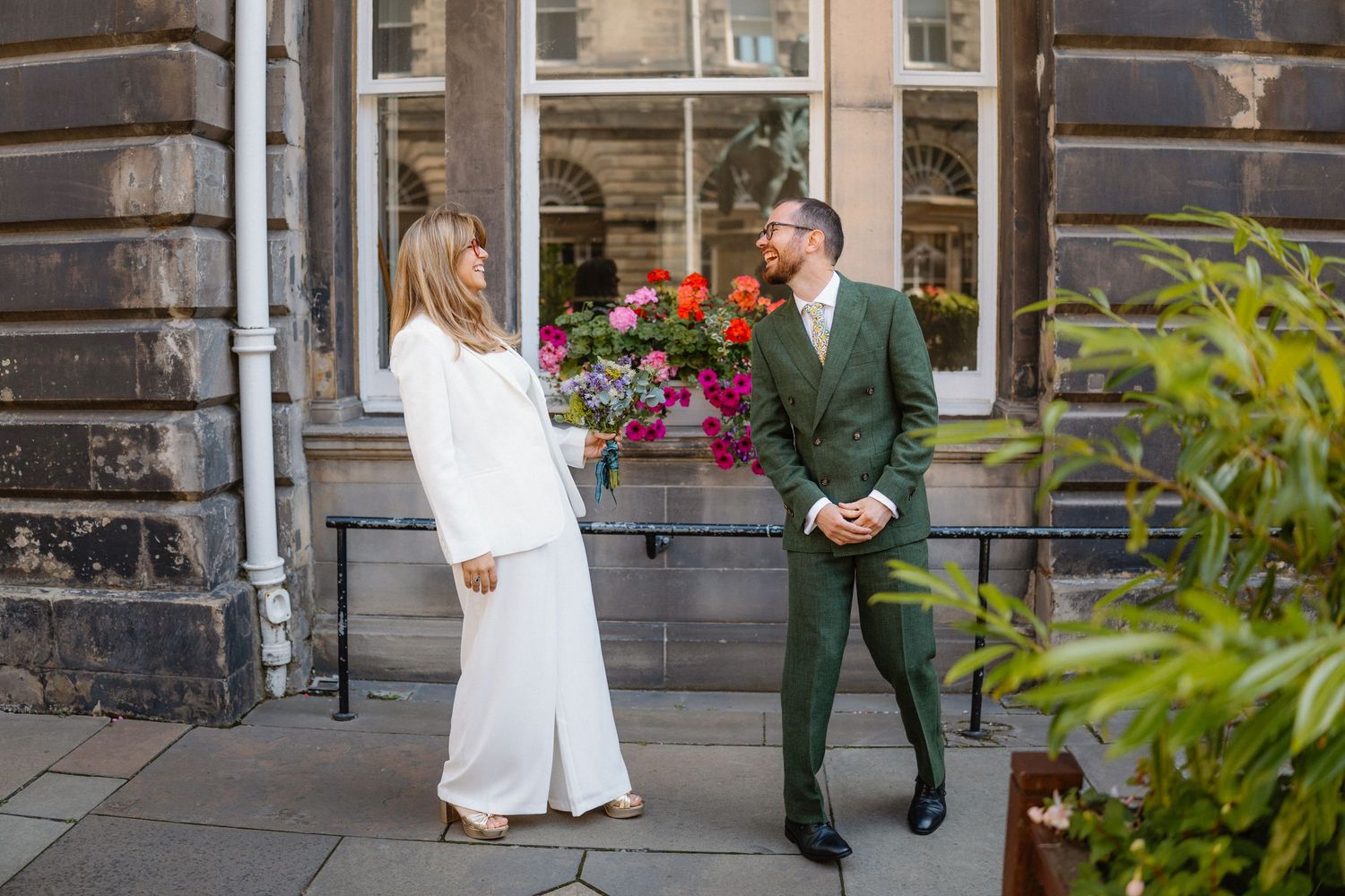 Wedding couple sharing a moment outside historic stone building with red flowers.