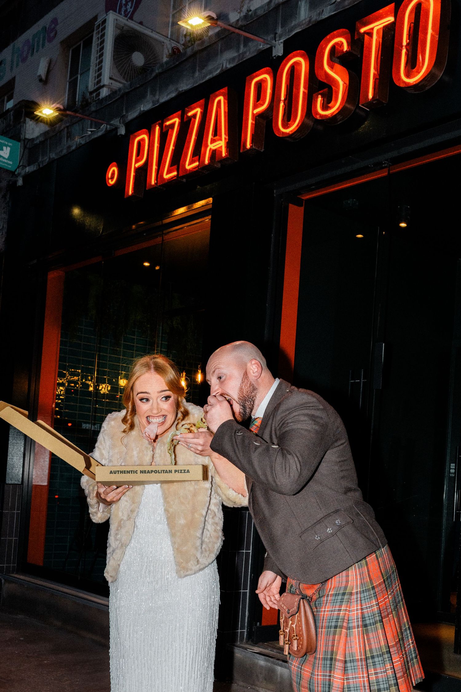 A couple playfully shares a long piece of bread outside a pizza restaurant with glowing red neon signs.