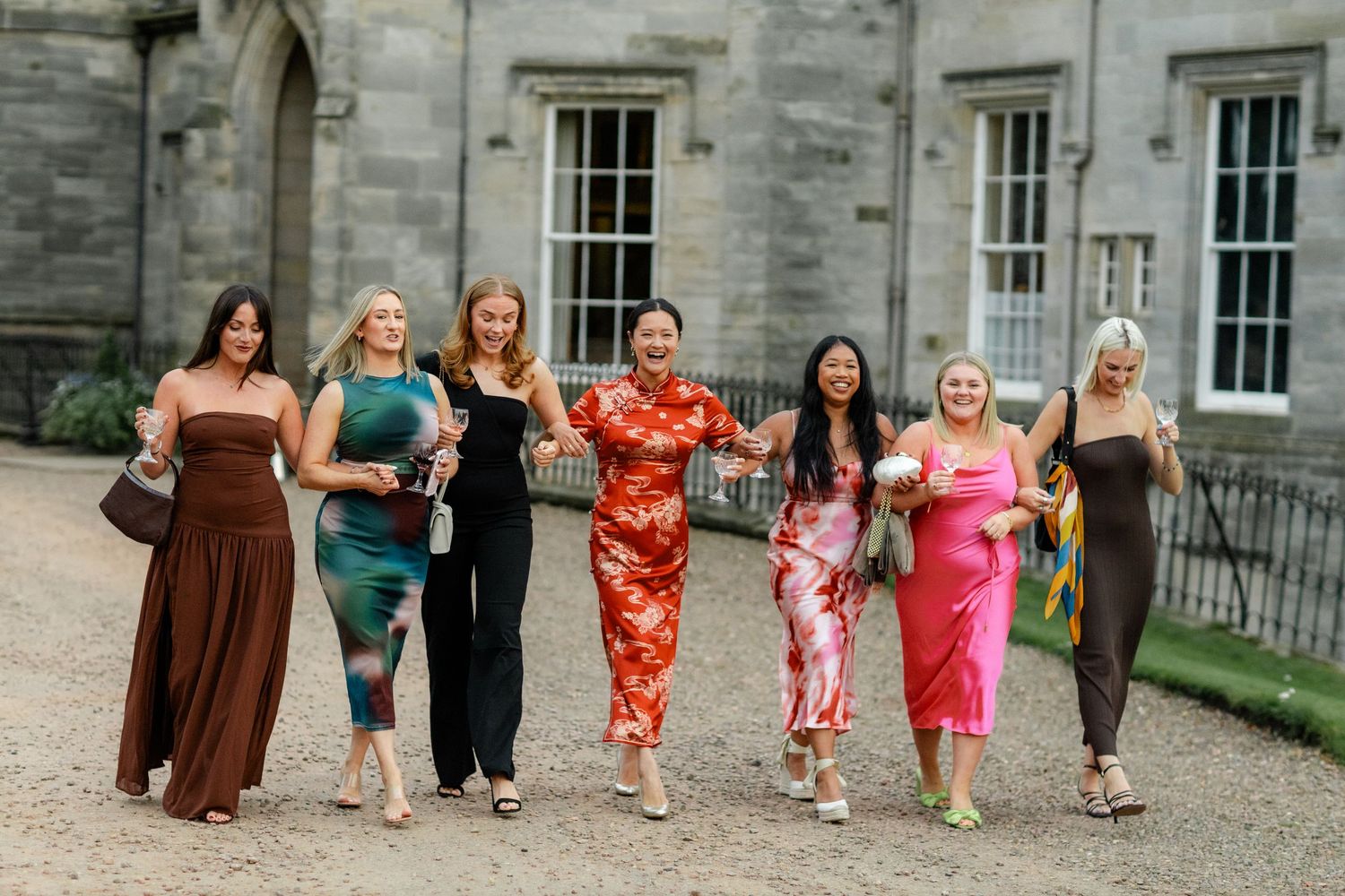 A group in colorful formal dresses walking together on a gravel path near a stone building.