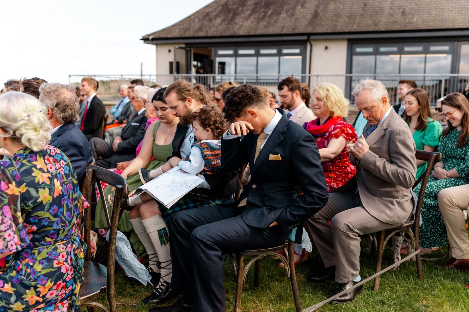 Wedding guests seated in rows of chairs share laughter during outdoor ceremony.