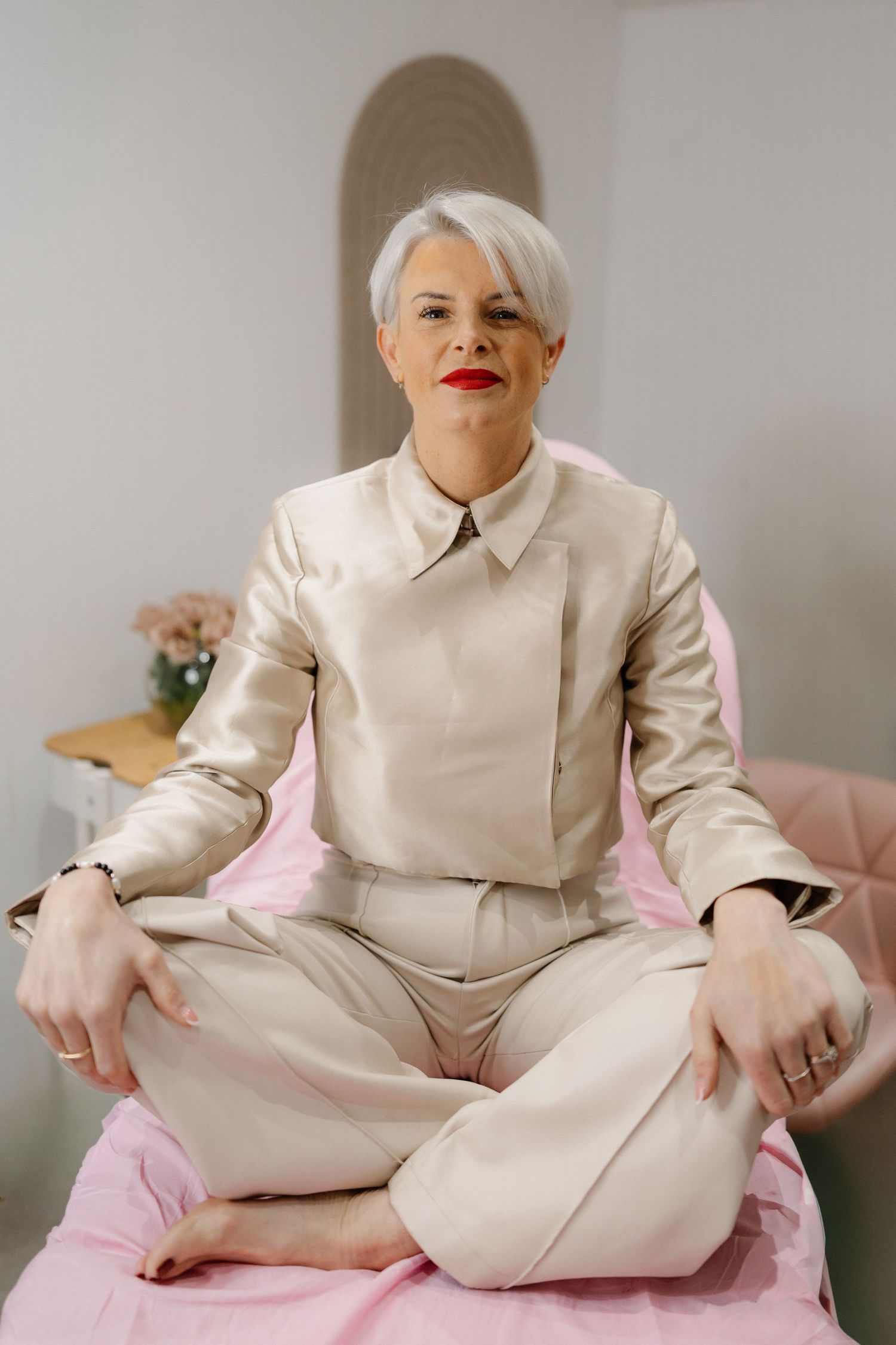 A person with platinum blonde hair wearing a cream colored suit sits cross-legged on a pink cushion.