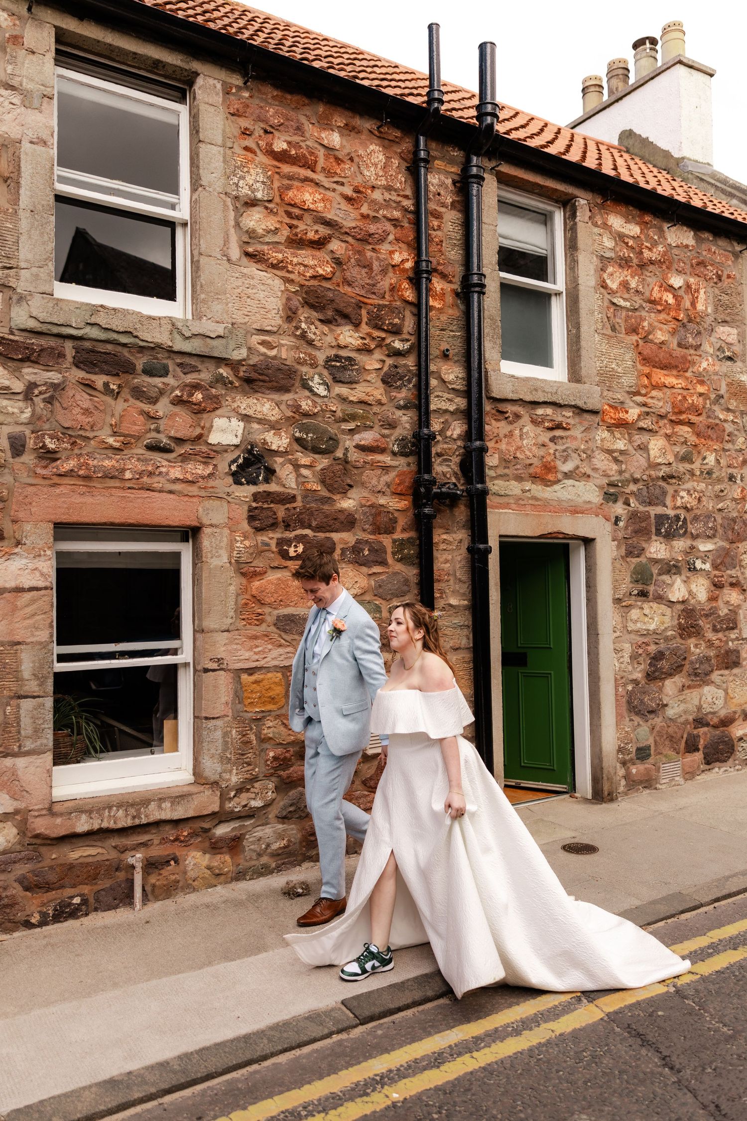 Wedding couple walking along rustic stone building with orange tiled roof.