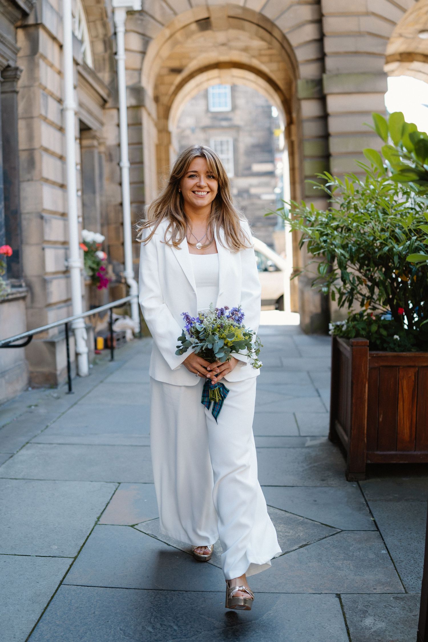 A bride in a flowing white dress and sweater holds a bouquet of blue flowers while walking under stone archways.
