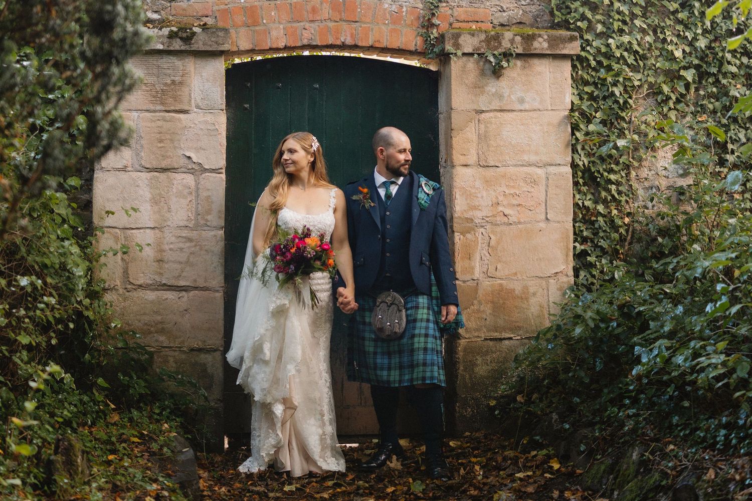 A couple in wedding attire poses against a rustic stone wall with ivy and greenery in an outdoor garden setting.