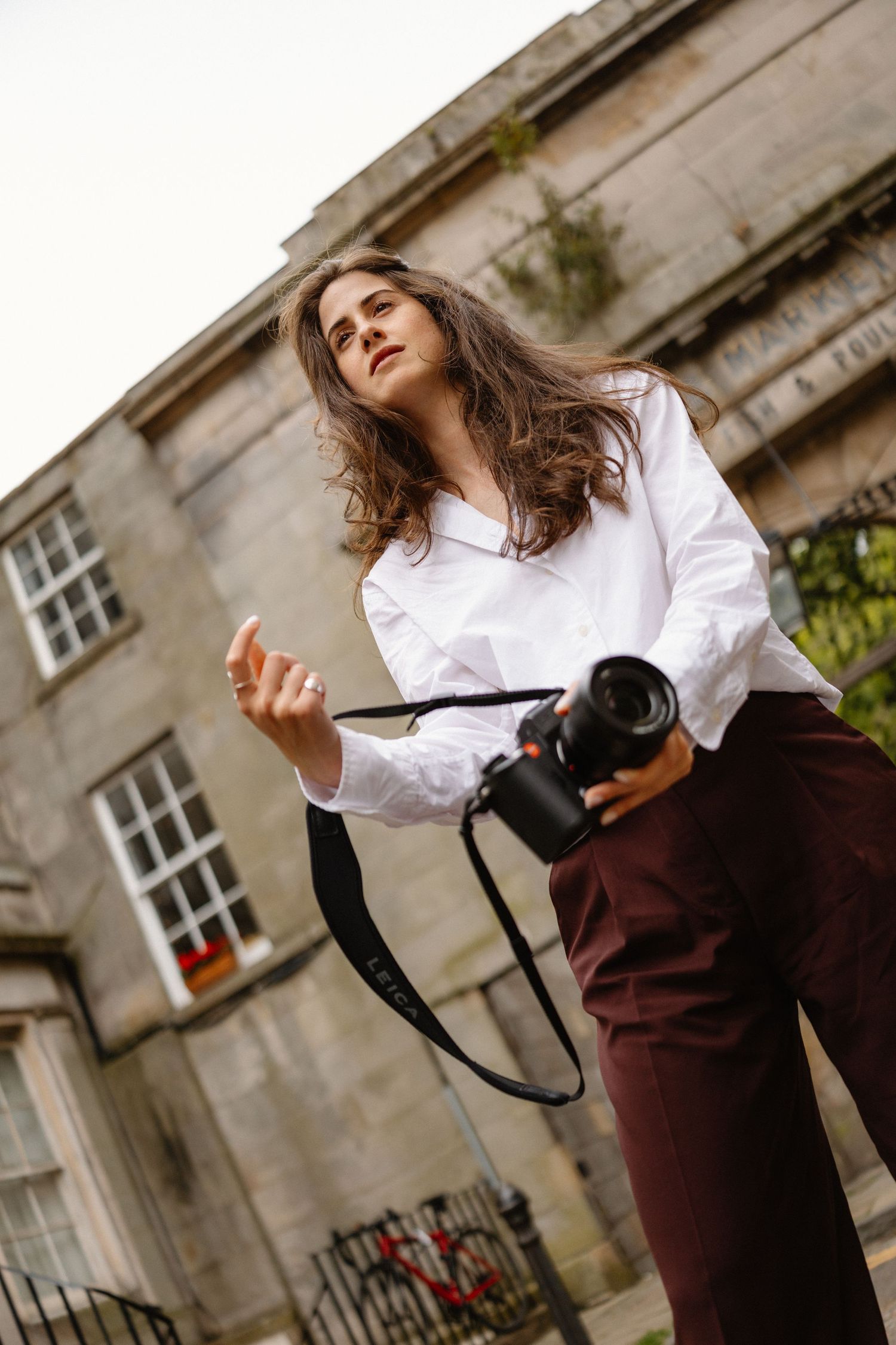 Photographer with camera stands near historic building in white shirt and burgundy pants.