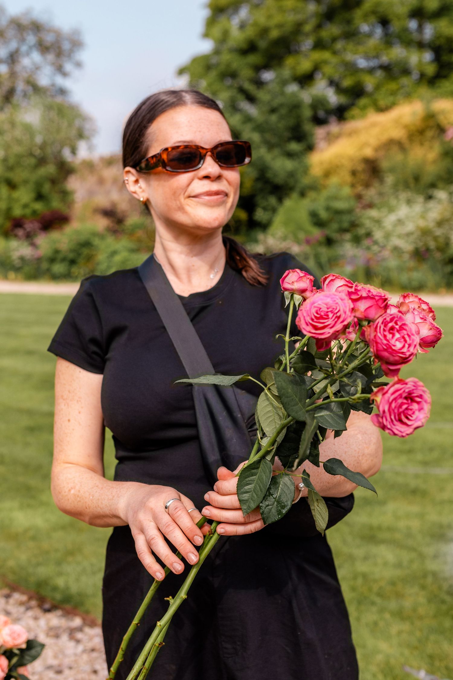 Someone in black holding fresh-cut pink roses in a sunny garden setting.