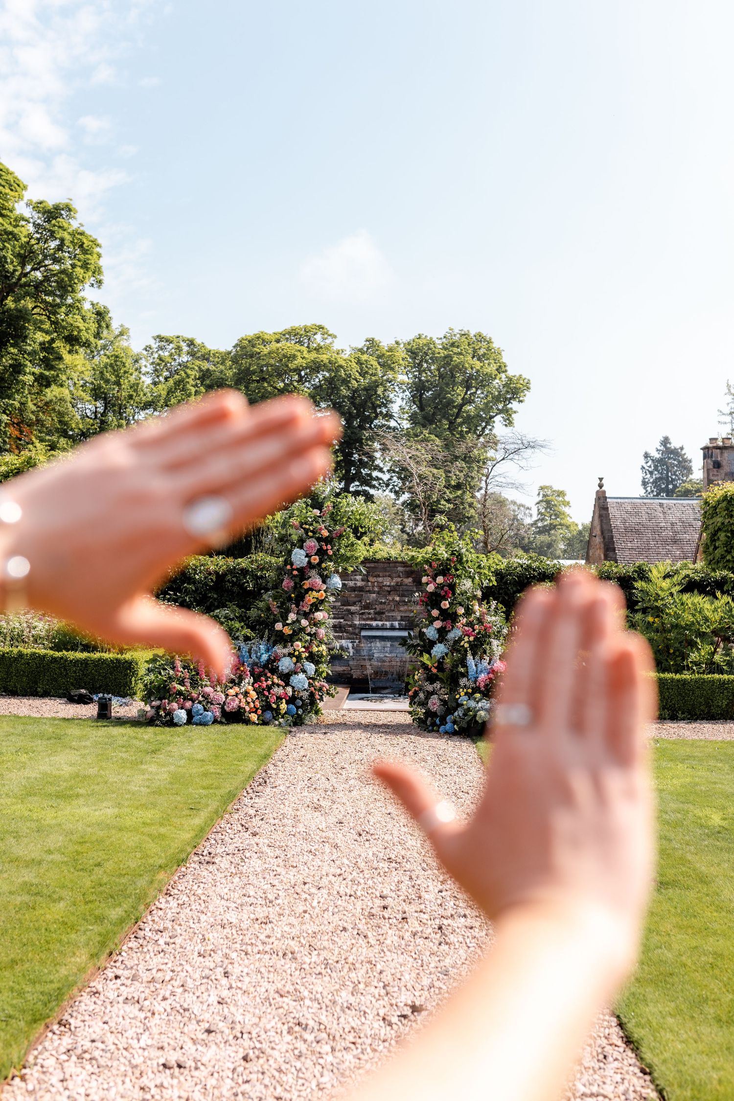 Hands creating a heart shape gesture against a sunny outdoor garden pathway background.