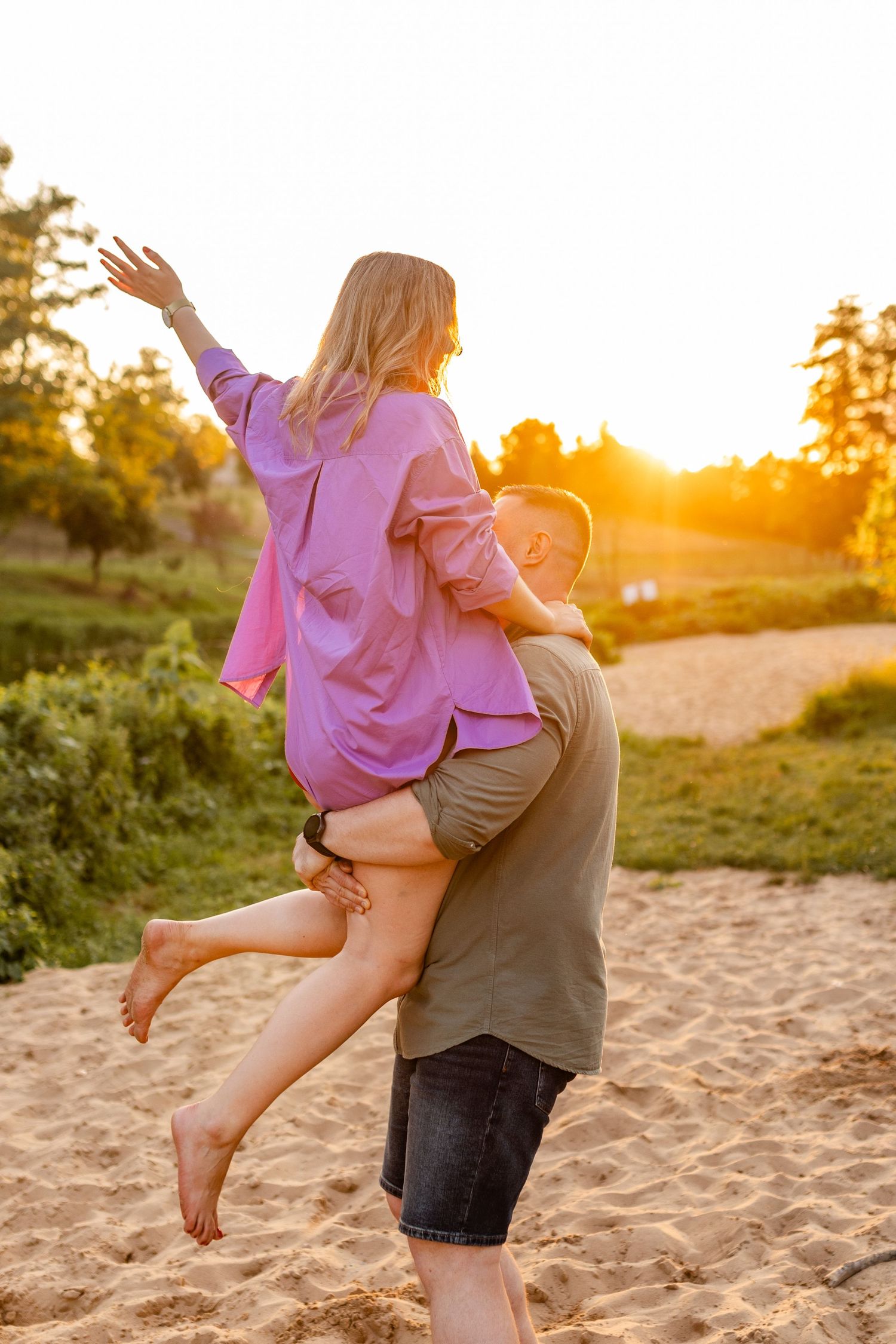 Playful couple poses on sandy beach at sunset with flowing purple clothing.