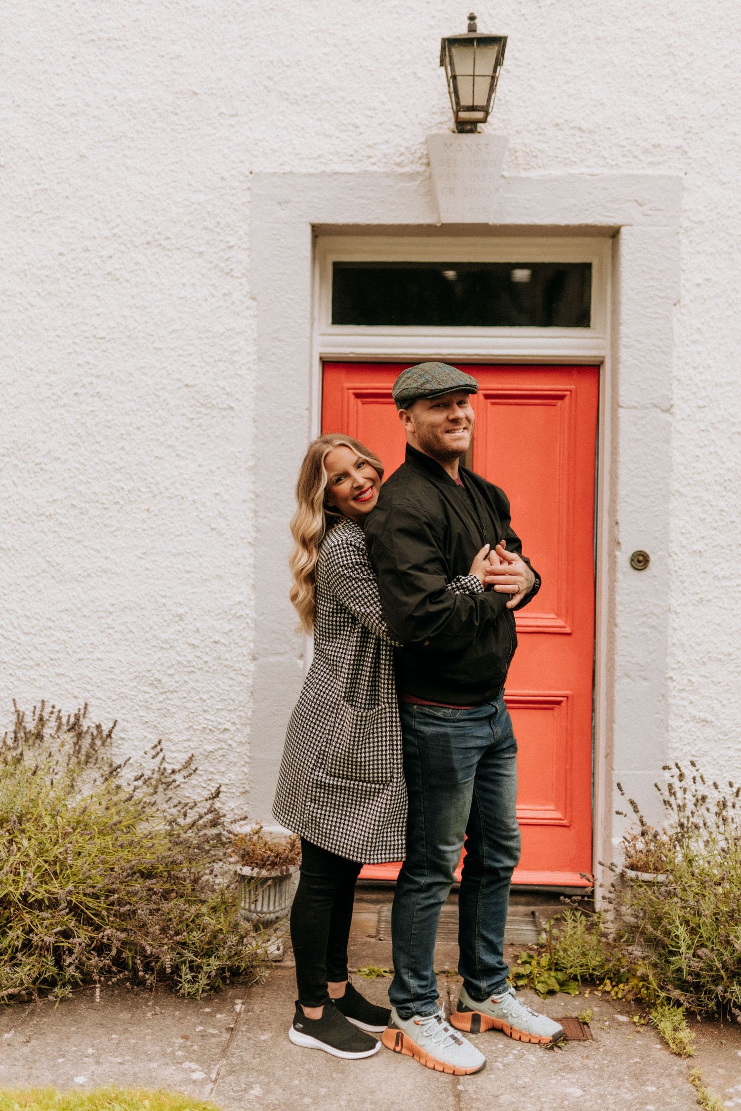 A couple embracing by a white building with a bright coral red door.
