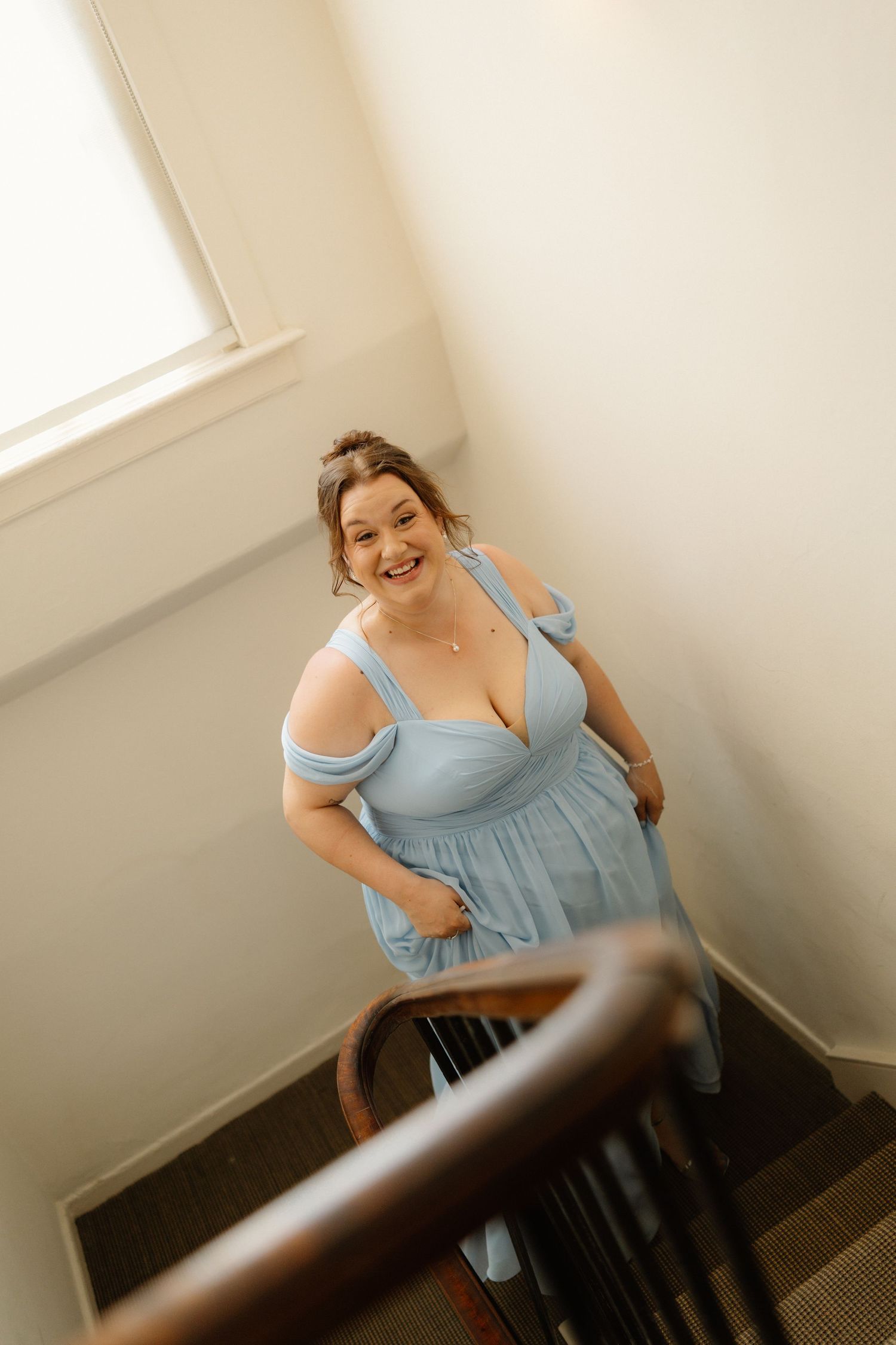 Smiling woman in a light blue off-shoulder dress ascending a wooden staircase in a bright interior.