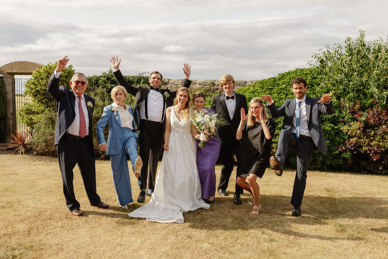 Wedding party celebrating outdoors, bride and groom with guests in formal attire, cheering and jumping with joy.