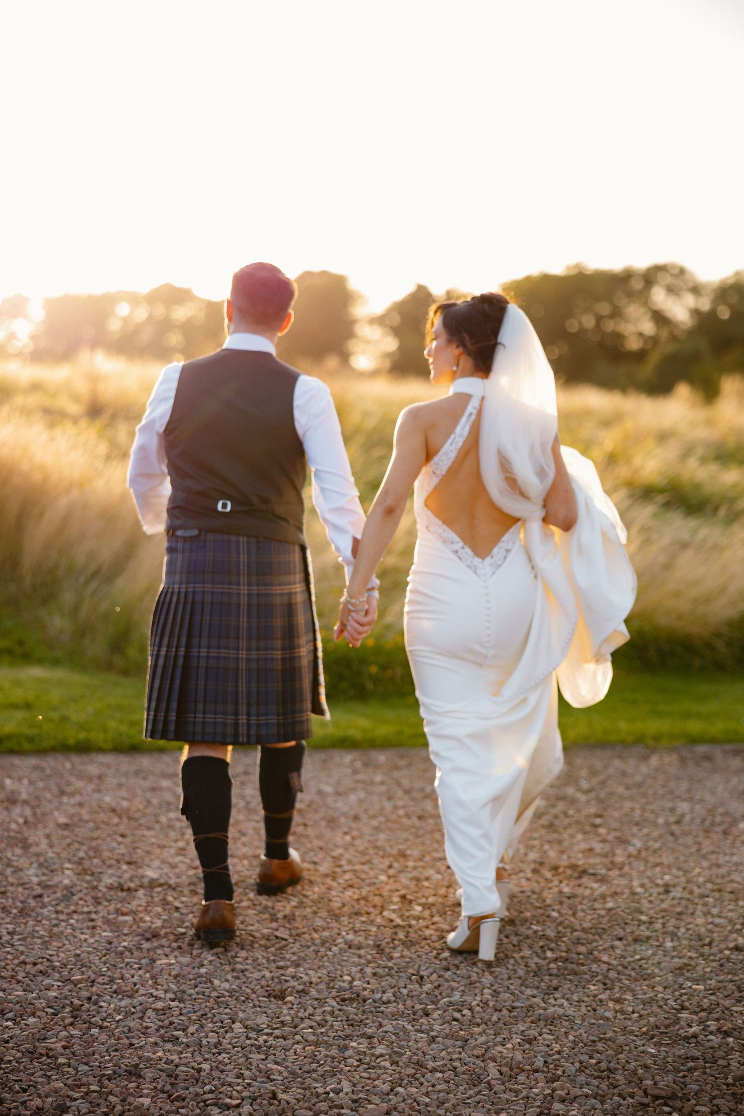 Bride and groom walk hand in hand at sunset, groom wearing traditional Scottish kilt on gravel path.