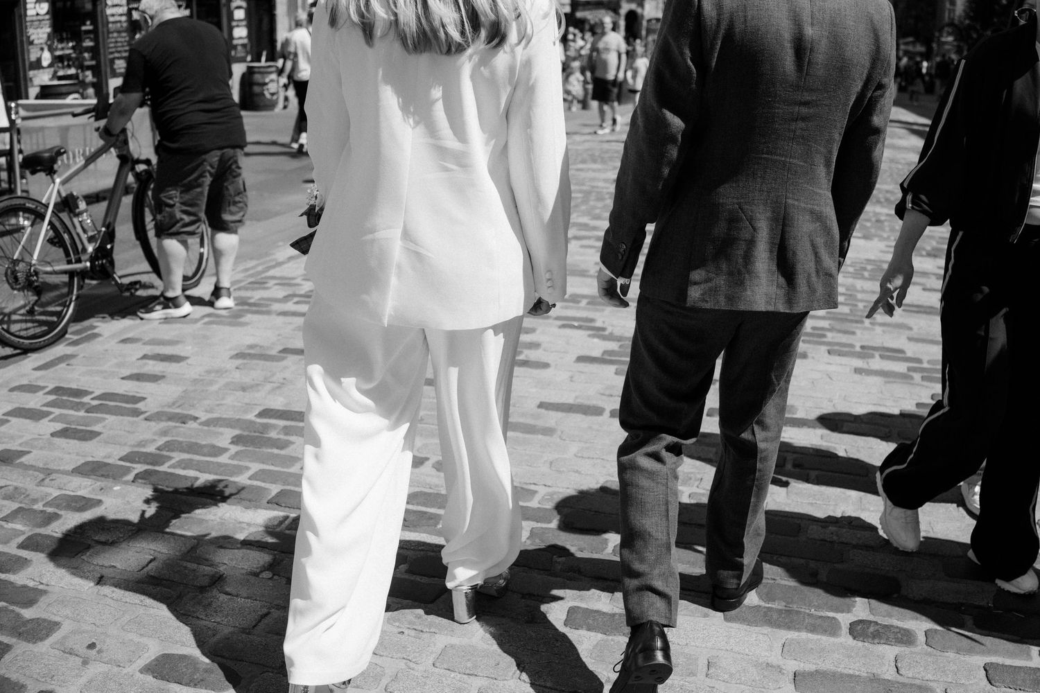 Black and white photo of a bride in white suit and groom walking on cobblestone streets.